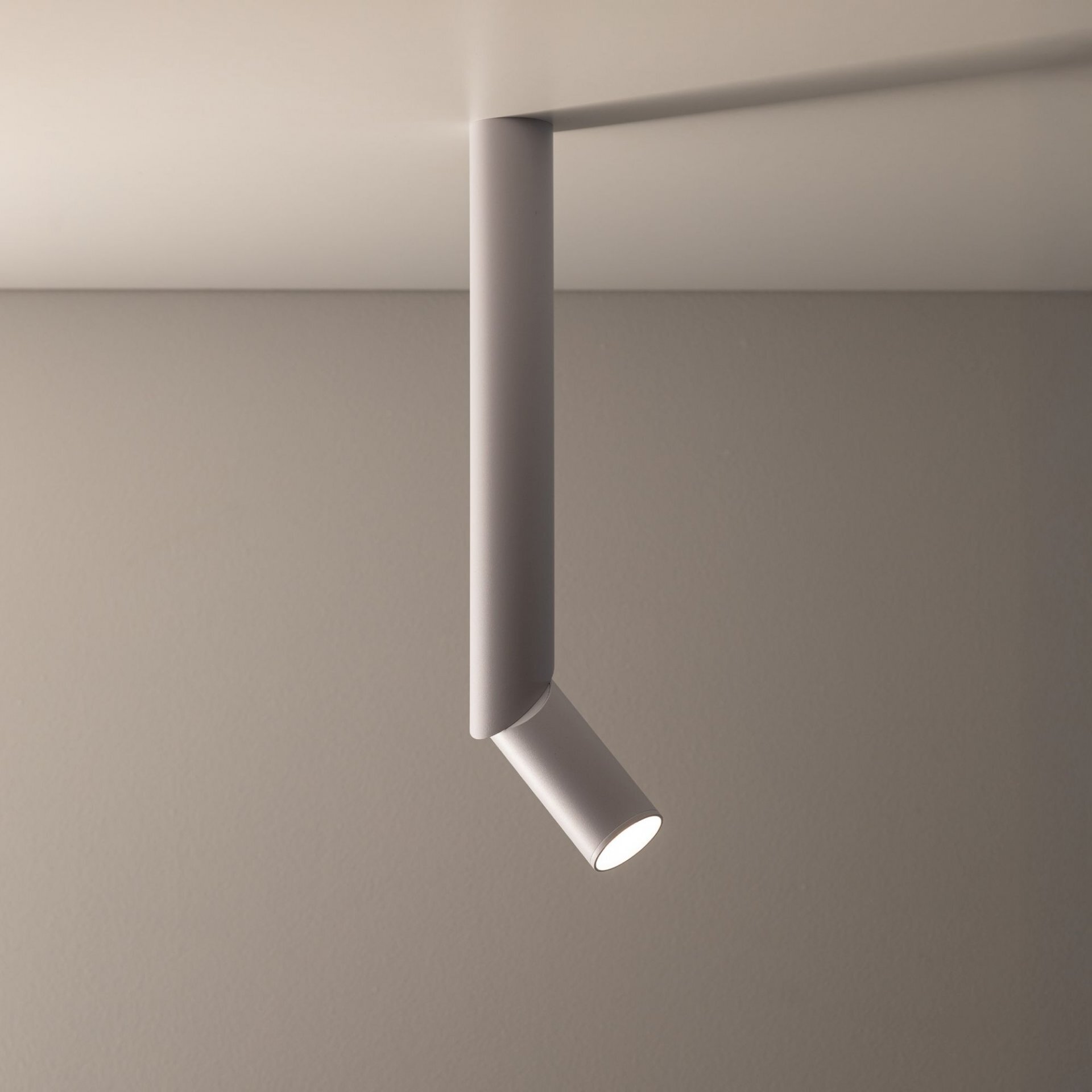 Biała lampa sufitowa w kształcie tuby zamontowana na suficie, z możliwością regulacji kierunku światła. Ściana w tle jest w neutralnym kolorze.