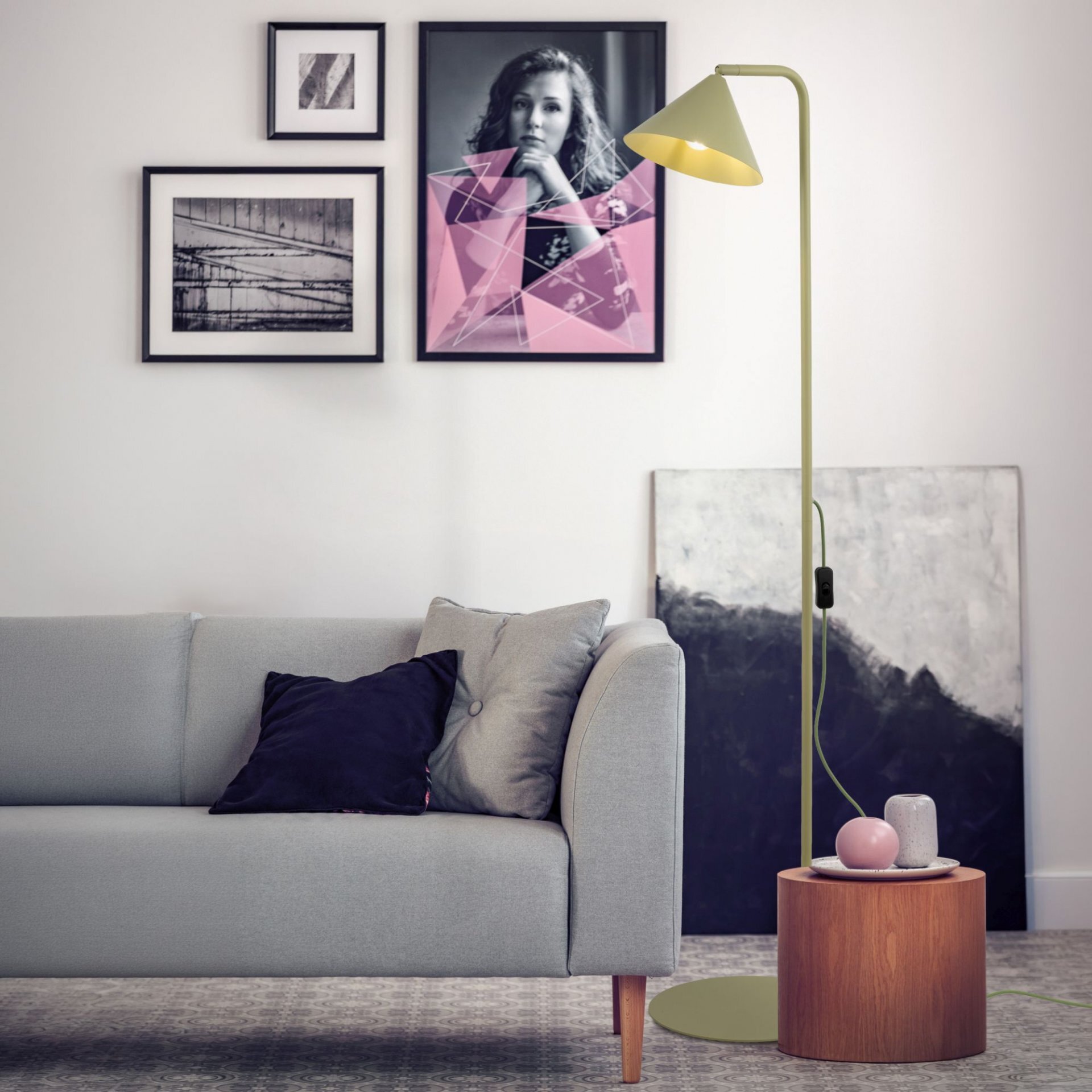 W salonie znajduje się nowoczesna lampa podłogowa w kolorze pistacjowym, stojąca obok szarej sofy. Na ścianie wiszą trzy obrazy, a na drewnianym stoliku leżą dekoracyjne przedmioty.