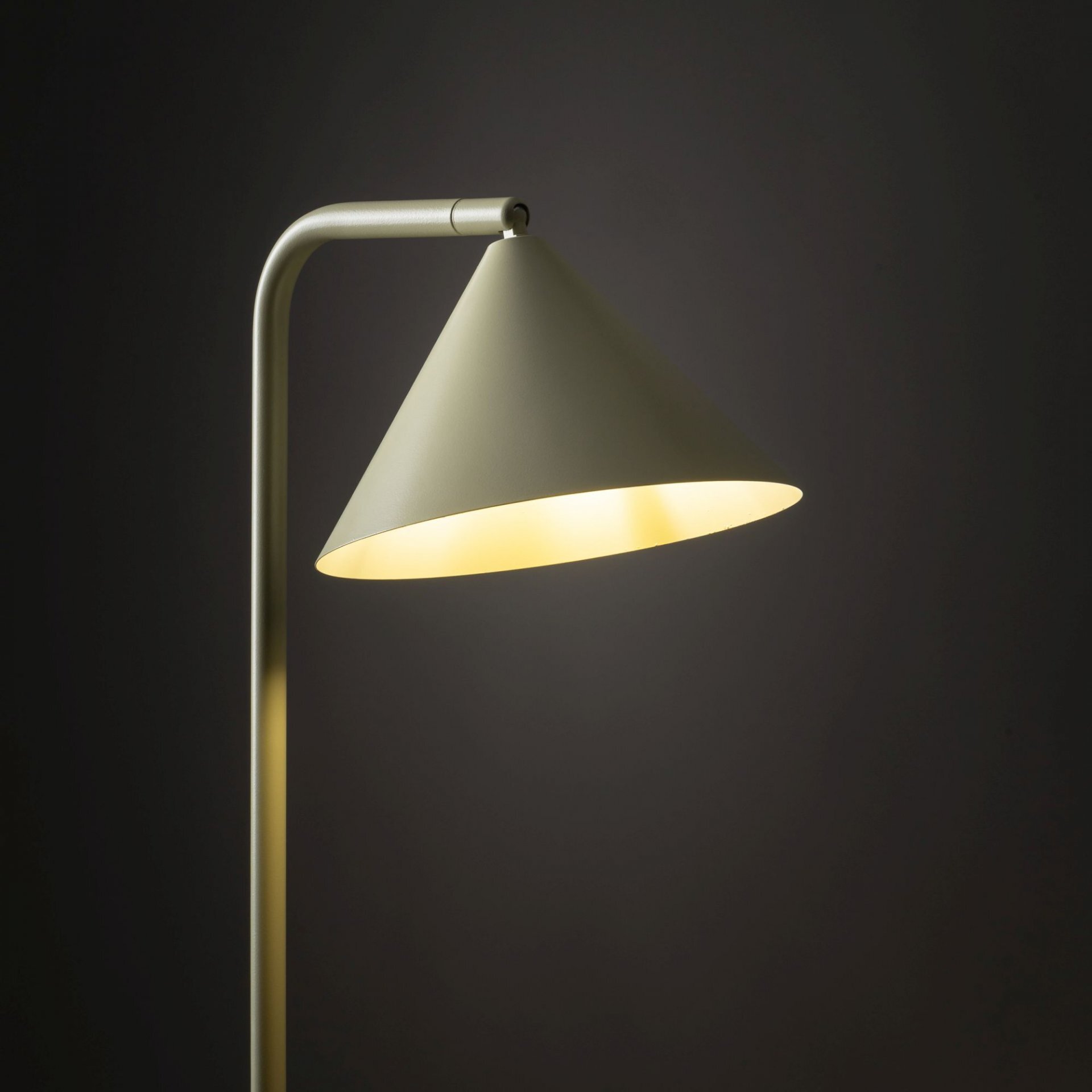 Lampa podłogowa o stożkowym kloszu w kolorze pistacjowym, emitująca ciepłe światło. Tło jest ciemne, co podkreśla jasność lampy.