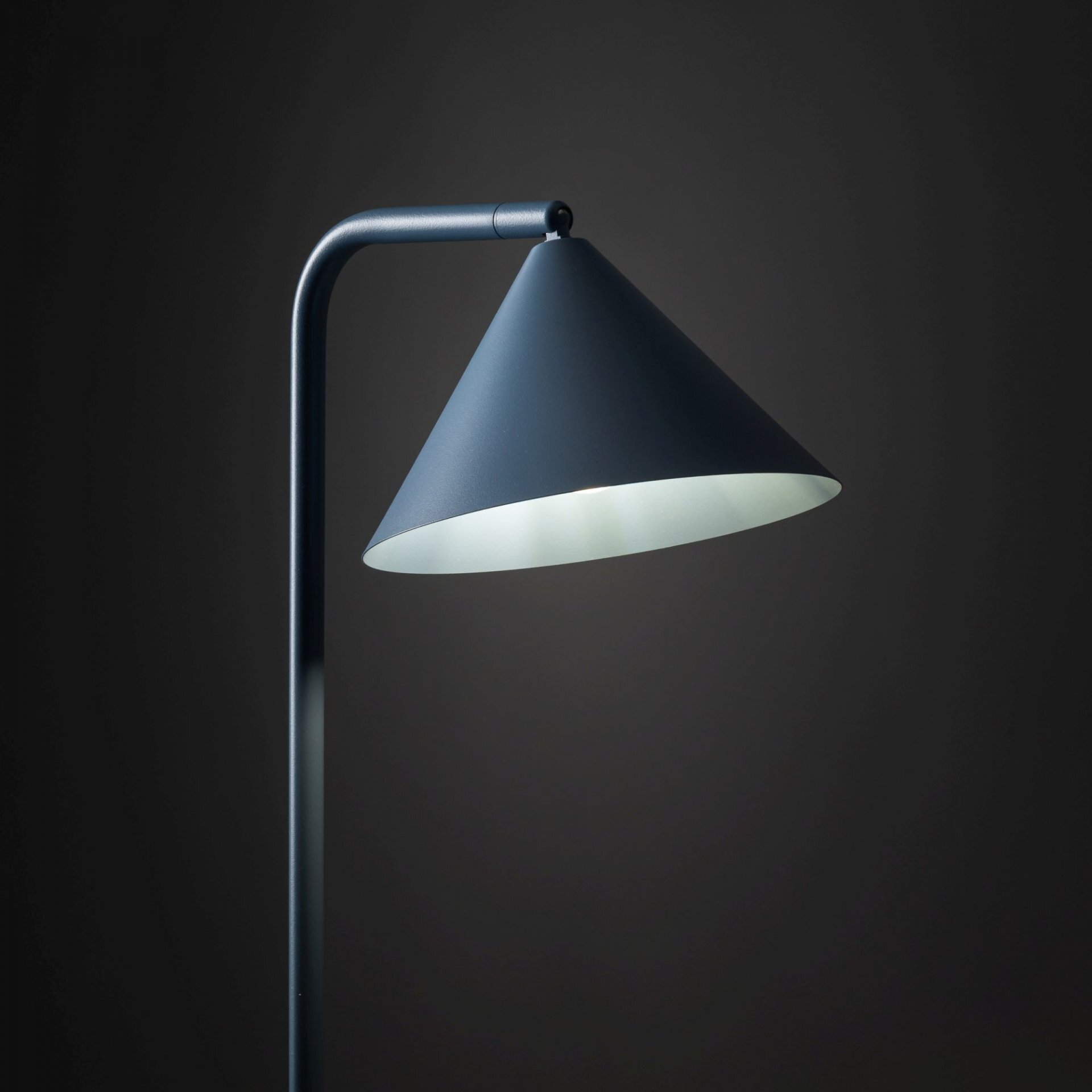 Lampa podłogowa o stożkowym kloszu w kolorze granatowym, z metalowym ramieniem na ciemnym tle.