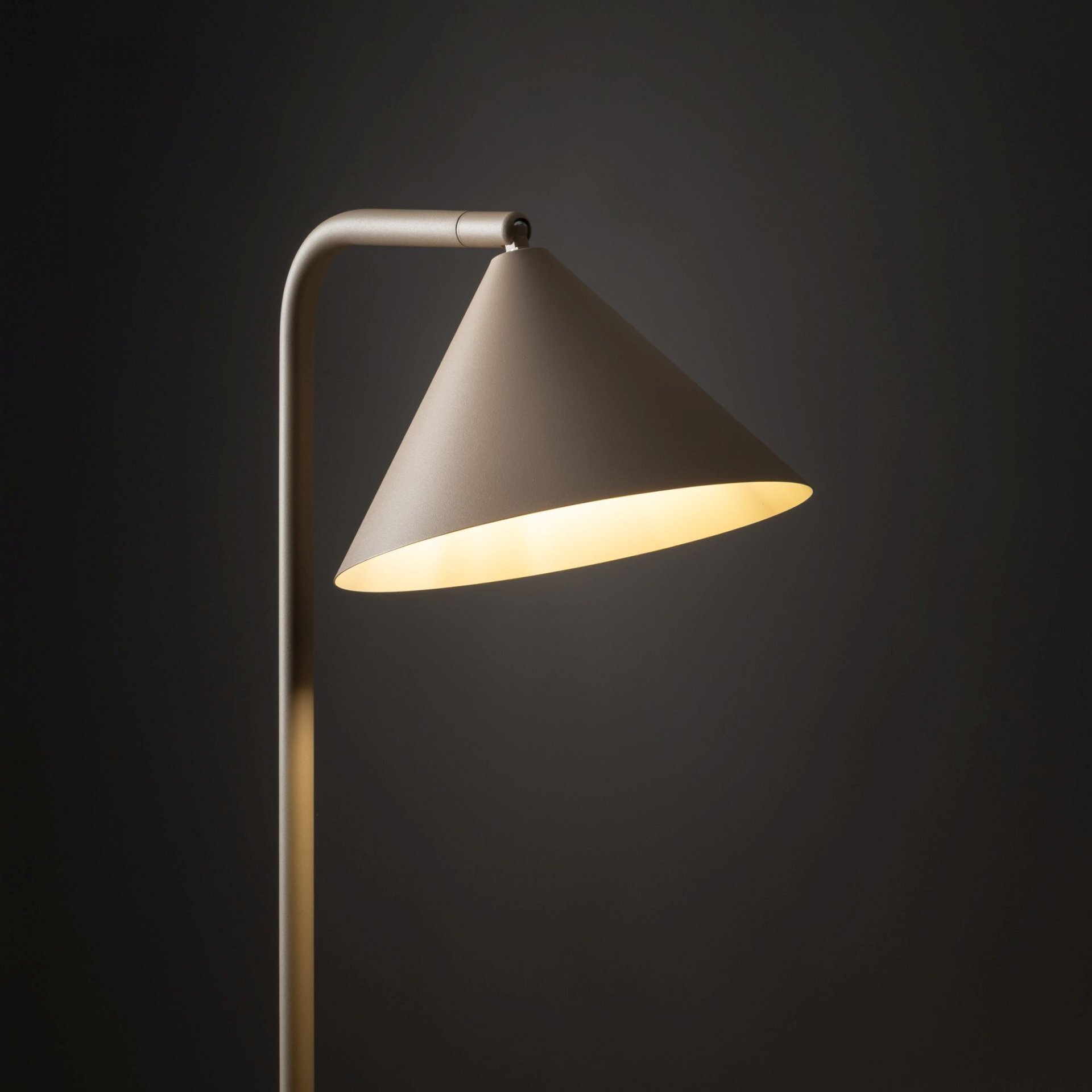 Lampa podłogowa z metalowym, stożkowym kloszem w kolorze beżowym, emitująca ciepłe światło. Tło jest ciemne, co podkreśla elegancki design lampy.