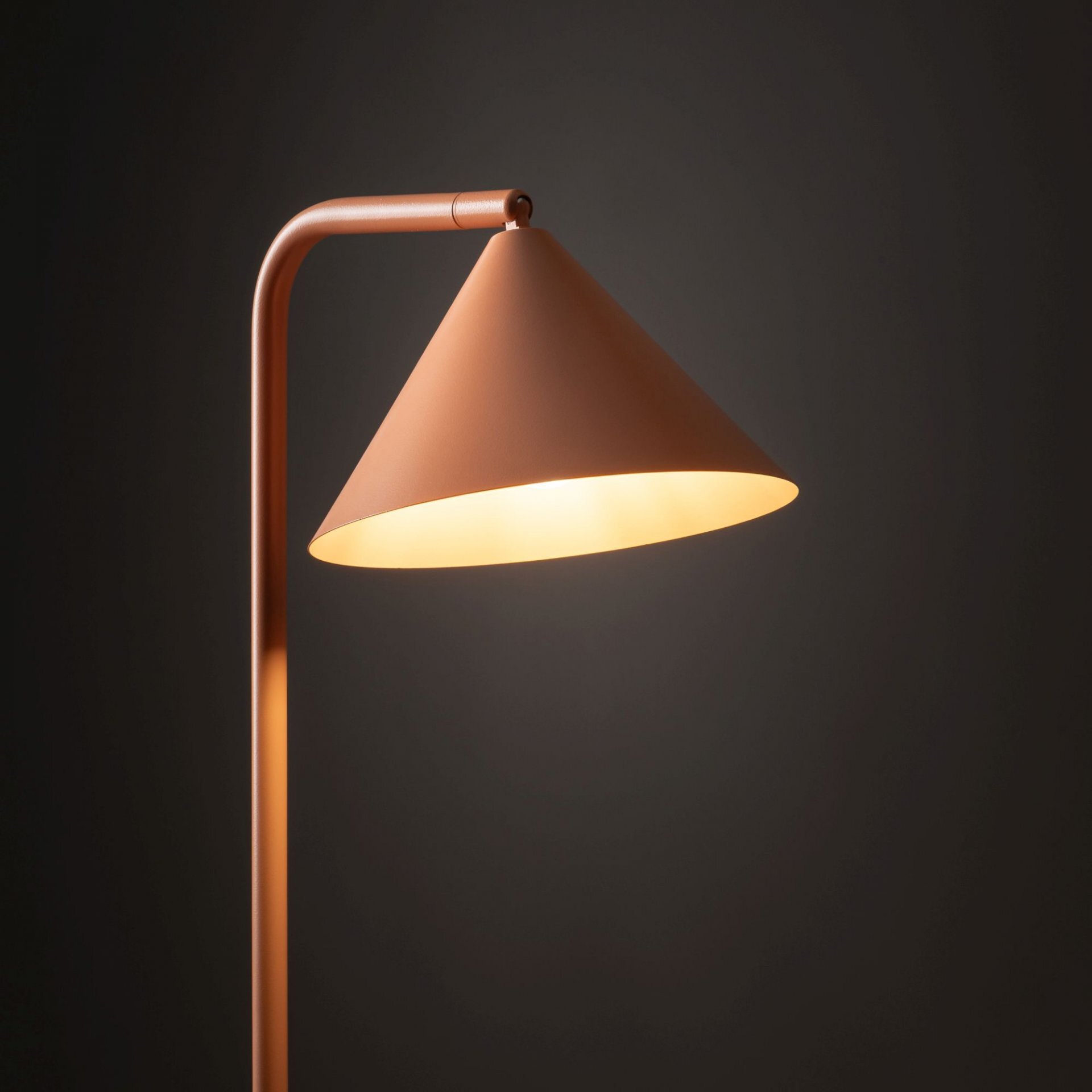 Lampa podłogowa o stożkowym kloszu w kolorze łososiowym, emitująca ciepłe światło. Tło jest ciemne, co podkreśla nowoczesny design lampy.