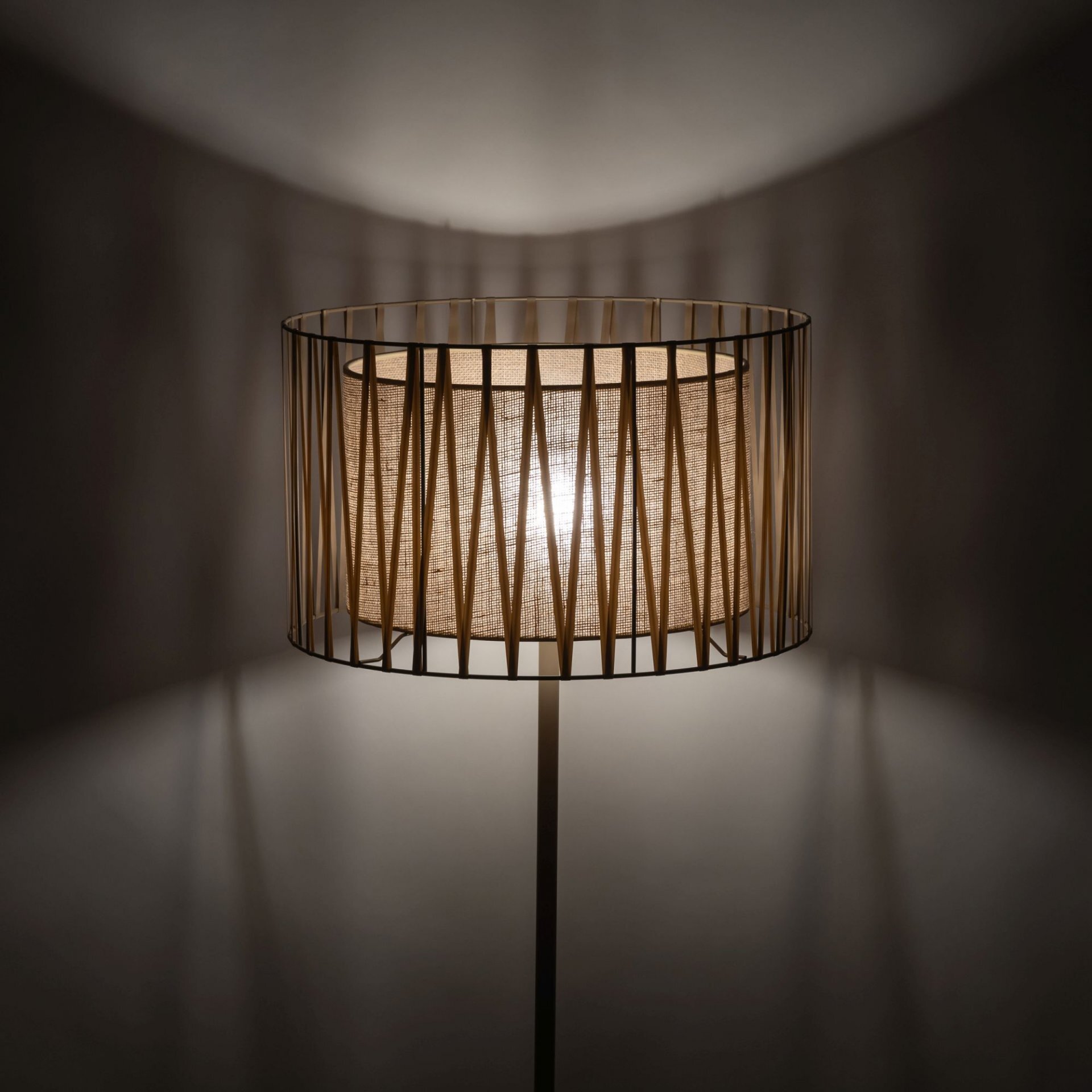 Lampa podłogowa z abażurem w naturalnym kolorze, otoczona metalowymi prętami tworzącymi geometryczny wzór. Światło lampy rzuca cienie na ścianę, tworząc efekt dekoracyjny.