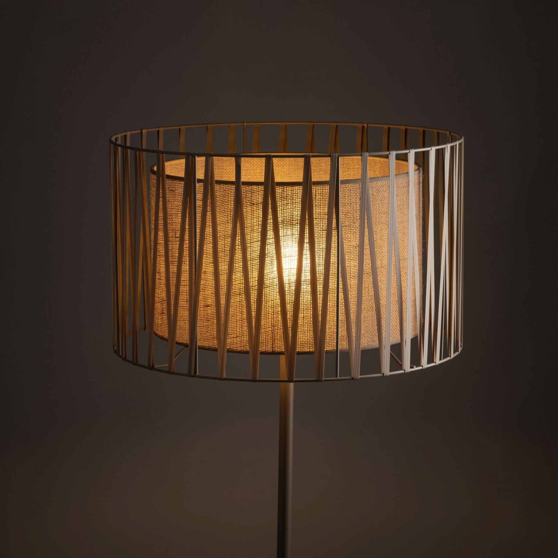 Lampa podłogowa z abażurem wykonanym z jasnego materiału, otoczona metalową konstrukcją o geometrycznym wzorze. Światło wewnątrz lampy tworzy ciepłą, przytulną atmosferę.
