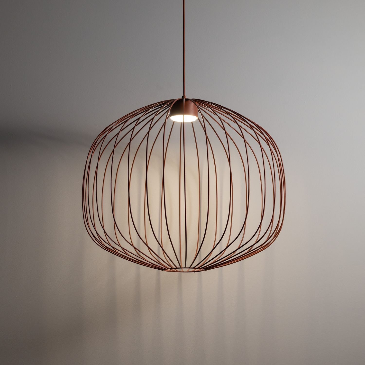 Lampa wisząca o nowoczesnym designie z metalowym kloszem w kształcie klatki, w kolorze ceglastym. Światło emituje z centralnego punktu, tworząc ciekawe cienie na ścianie.
