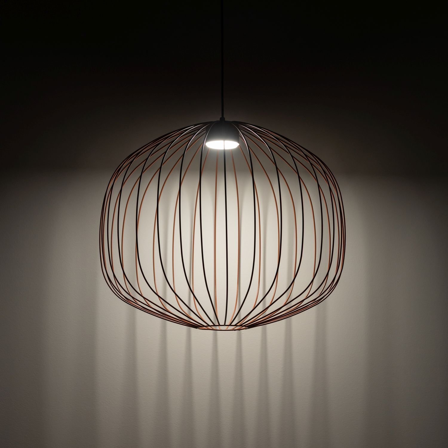 Na obrazie widoczna jest wisząca lampa z metalowym kloszem w kształcie kuli, przez który przechodzi światło, tworząc cienie na ścianie. Lampa jest włączona, emitując jasne światło skierowane w dół.