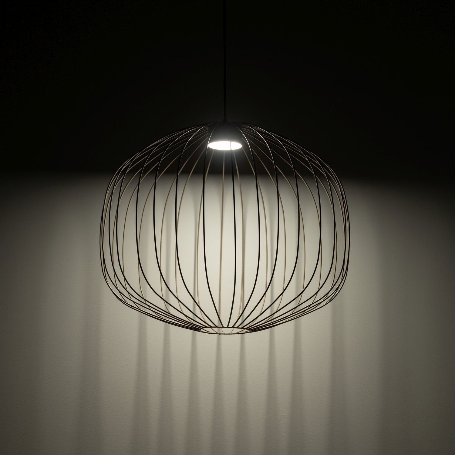 Lampa wisząca o ażurowej konstrukcji z metalowych prętów, emitująca światło z centralnego punktu. Światło tworzy cienie na ścianie, podkreślając geometryczny wzór lampy.