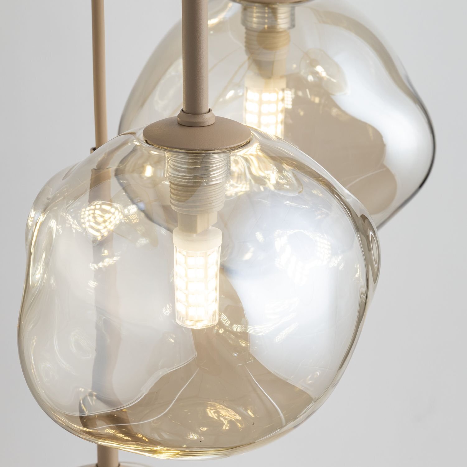 Zdjęcie przedstawia zbliżenie na szklane klosze lampy wiszącej z widocznymi żarówkami LED wewnątrz. Klosze mają nieregularny, organiczny kształt i są przezroczyste z delikatnym złotym odcieniem.