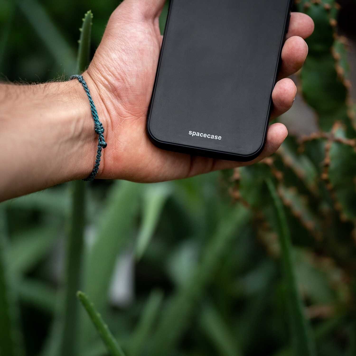 Osoba trzyma czarne etui na telefon z widocznym napisem \'spacecase\'. W tle znajdują się zielone rośliny.