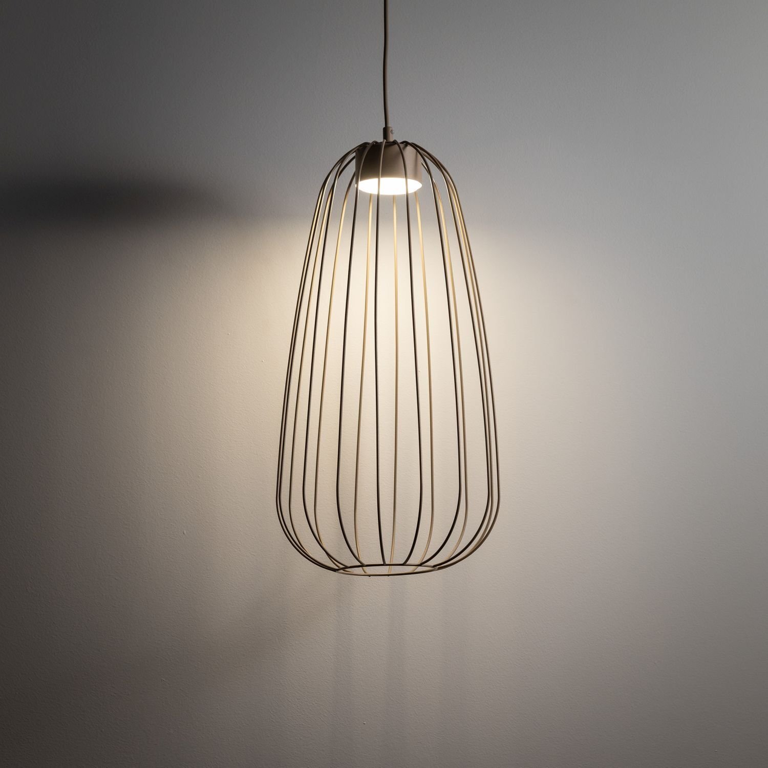 Lampa wisząca o nowoczesnym designie z metalowym kloszem w kształcie klatki, emitująca światło na ścianę. Lampa jest zawieszona na cienkim przewodzie.