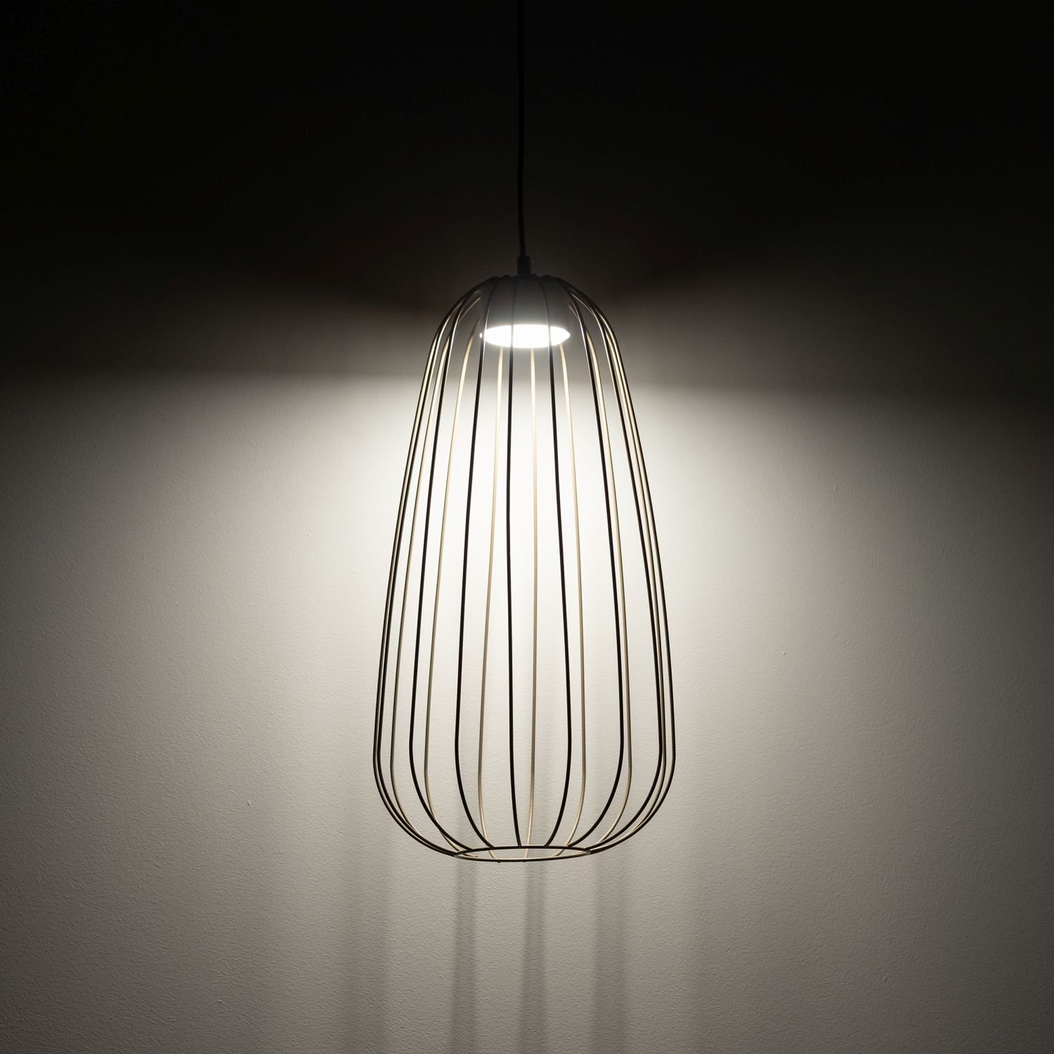 Lampa wisząca o nowoczesnym designie z metalowym kloszem w kształcie klatki, emitująca światło na ścianę. Światło tworzy delikatny efekt cieni na powierzchni.