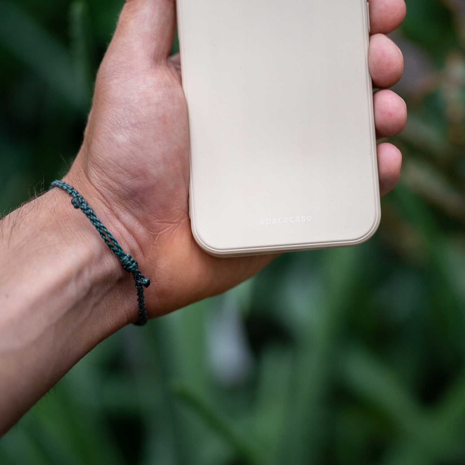 Osoba trzyma beżowe silikonowe etui na telefon z widocznym napisem \'spacecase\'. W tle znajdują się rozmyte zielone rośliny.