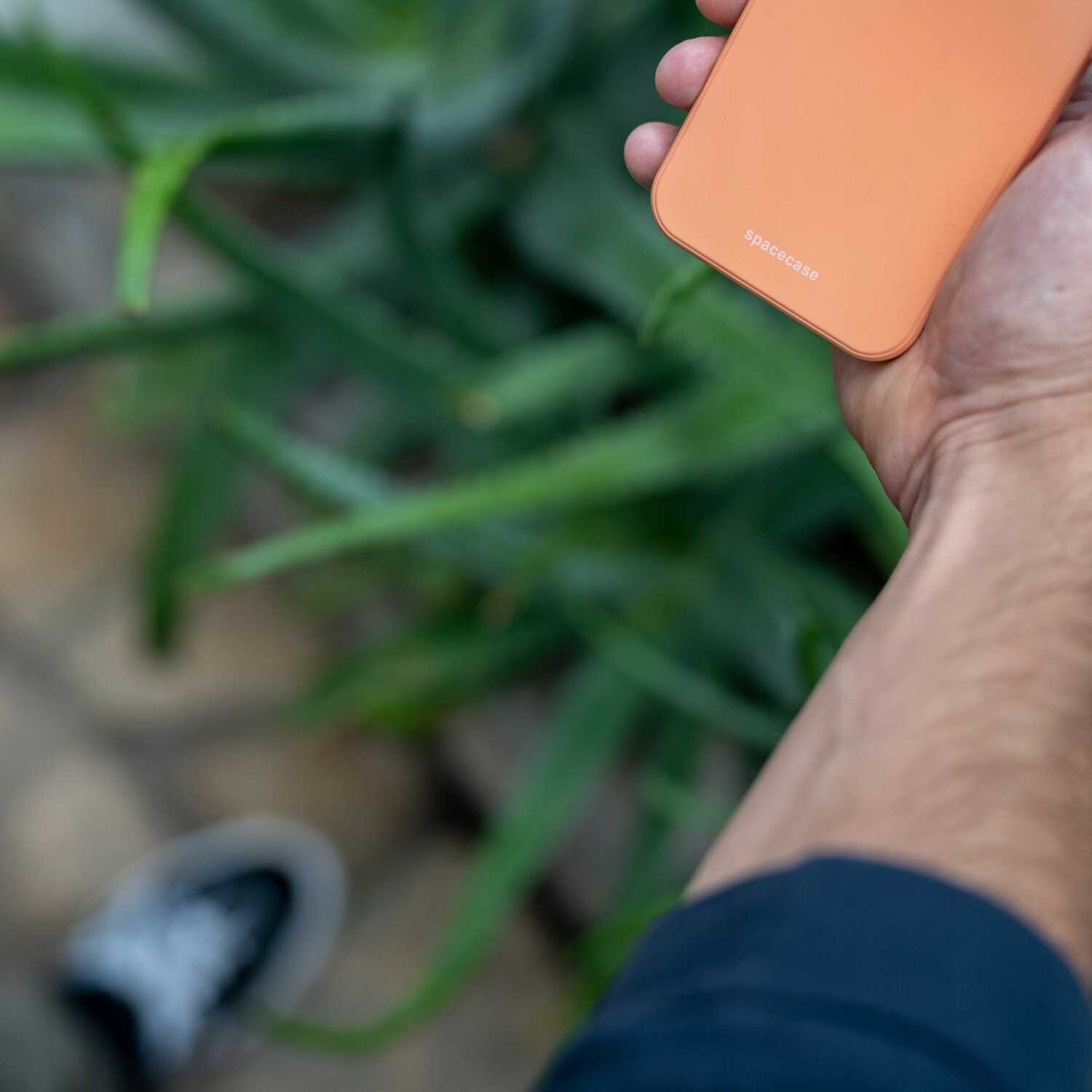 Osoba trzyma pomarańczowe silikonowe etui z napisem \'spacecase\'. W tle widoczne są zielone liście rośliny.