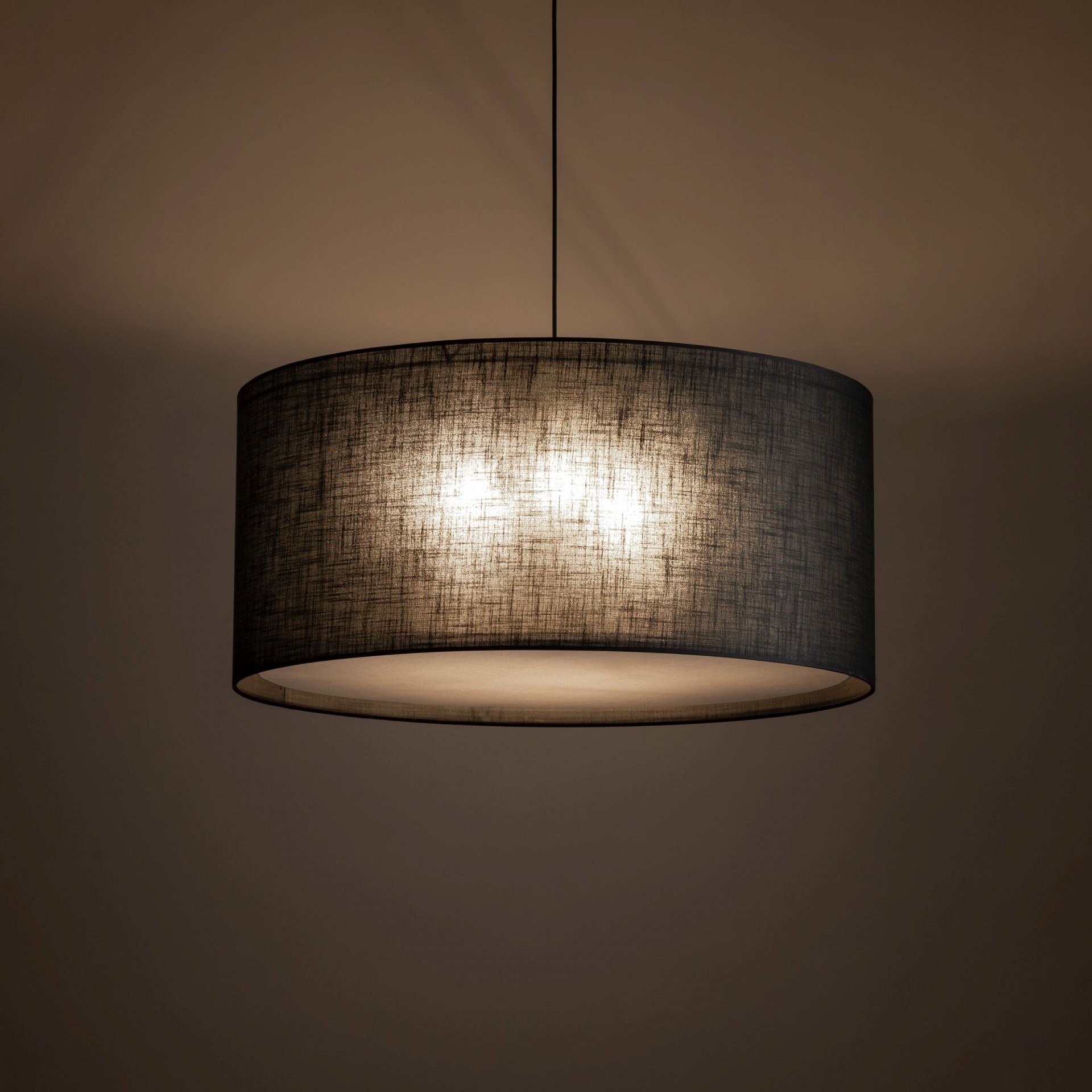Czarna, nowoczesna lampa wisząca z okrągłym abażurem, emitująca ciepłe światło. Lampa zawieszona jest na cienkim przewodzie, a jej powierzchnia ma teksturę przypominającą tkaninę.