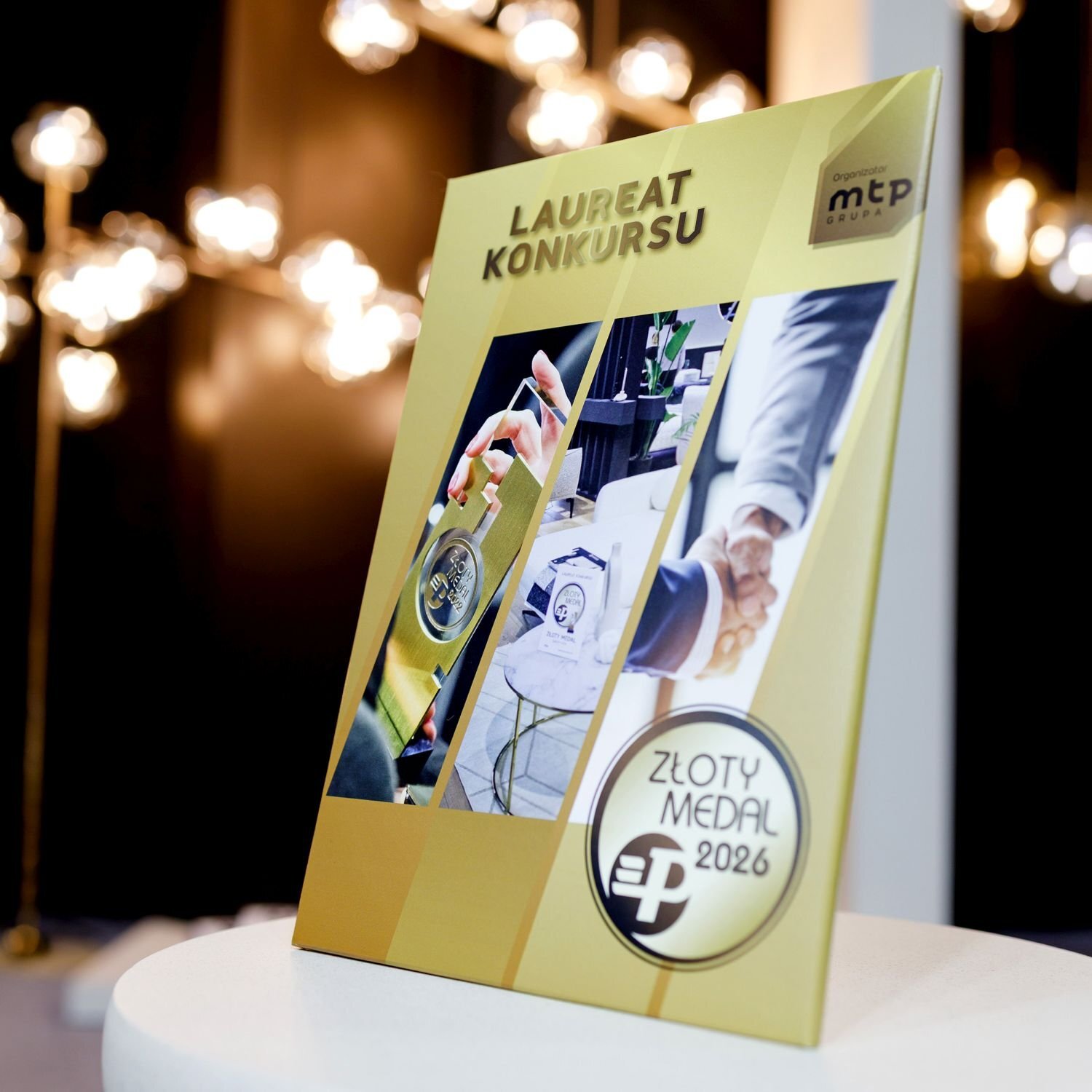 Na obrazie widoczna jest złota tablica z napisem \'LAUREAT KONKURSU\' oraz logo \'ZŁOTY MEDAL 2026\'. W tle znajdują się rozmyte światła lamp.