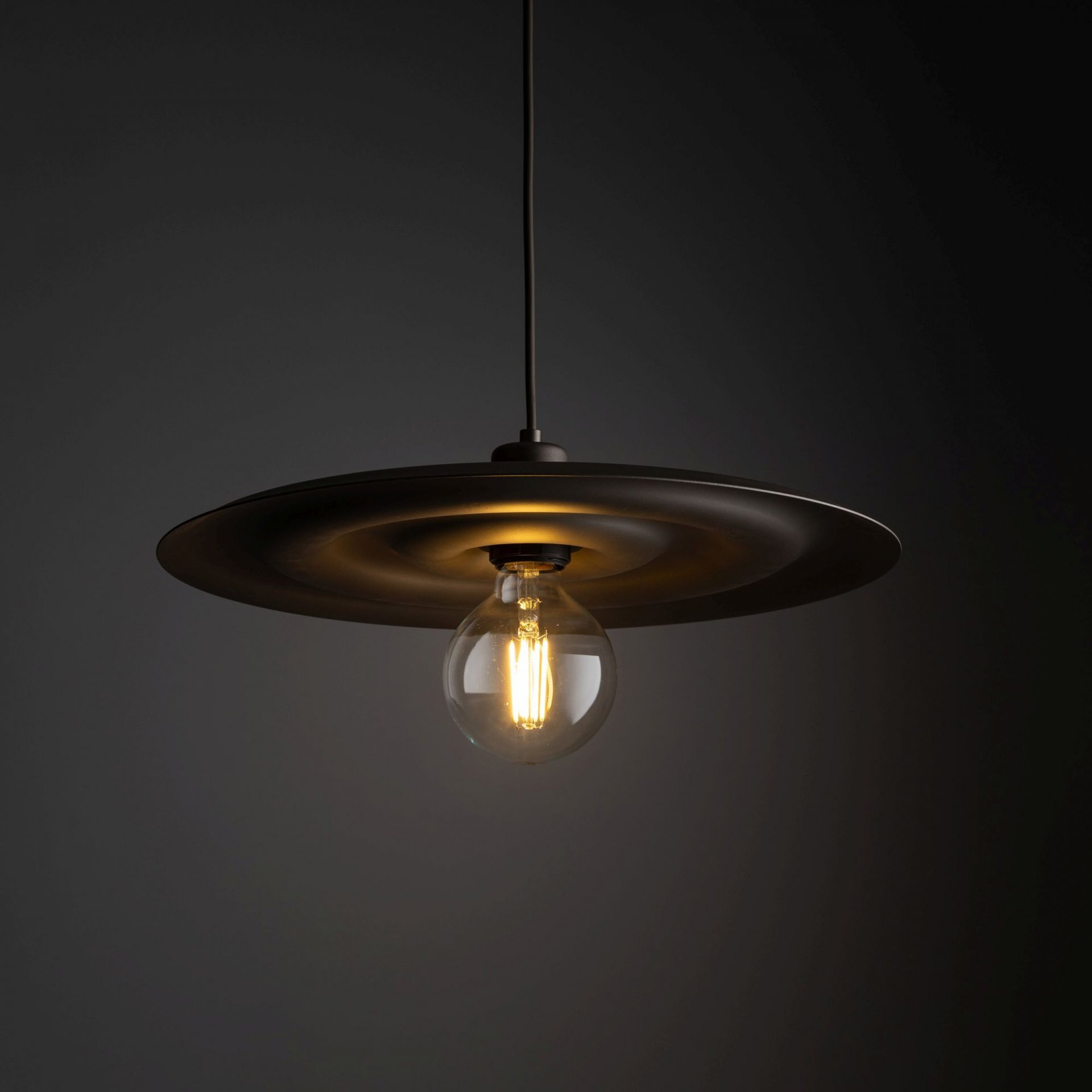 Na ciemnym tle wisi lampa z brązowym, okrągłym kloszem i widoczną żarówką w środku.