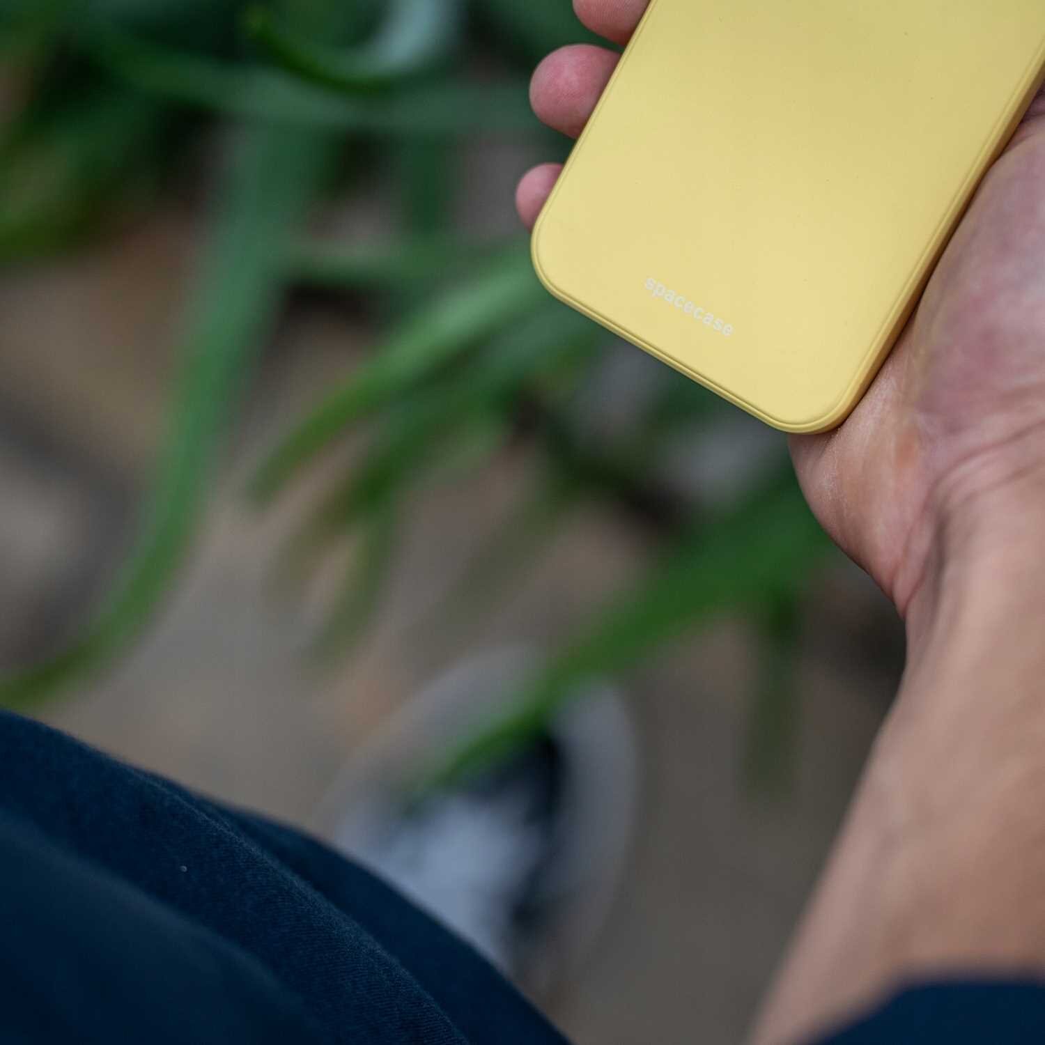 Osoba trzyma w ręku żółte silikonowe etui na telefon z napisem \'spacecase\'. W tle widoczne są rozmyte zielone liście.