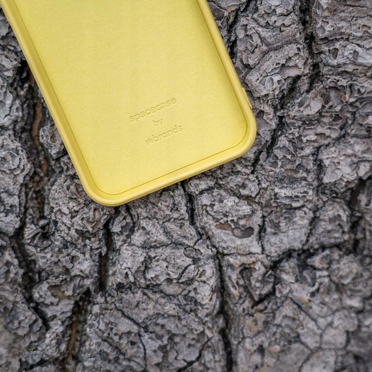 Żółte silikonowe etui leży na tle kory drzewa. Na etui widoczny jest napis: \'spacecase by nibrands\'.