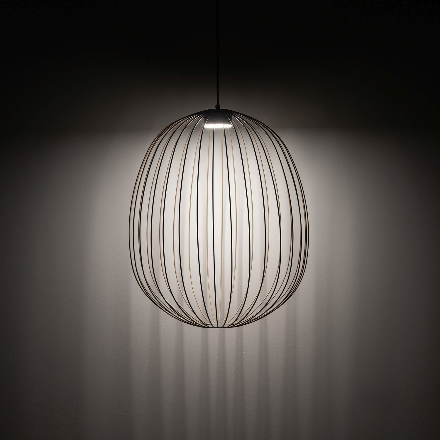 Na obrazie widoczna jest wisząca lampa o nowoczesnym designie, składająca się z metalowych prętów tworzących owalny kształt. Lampa emituje światło, które tworzy cienie na ścianie.