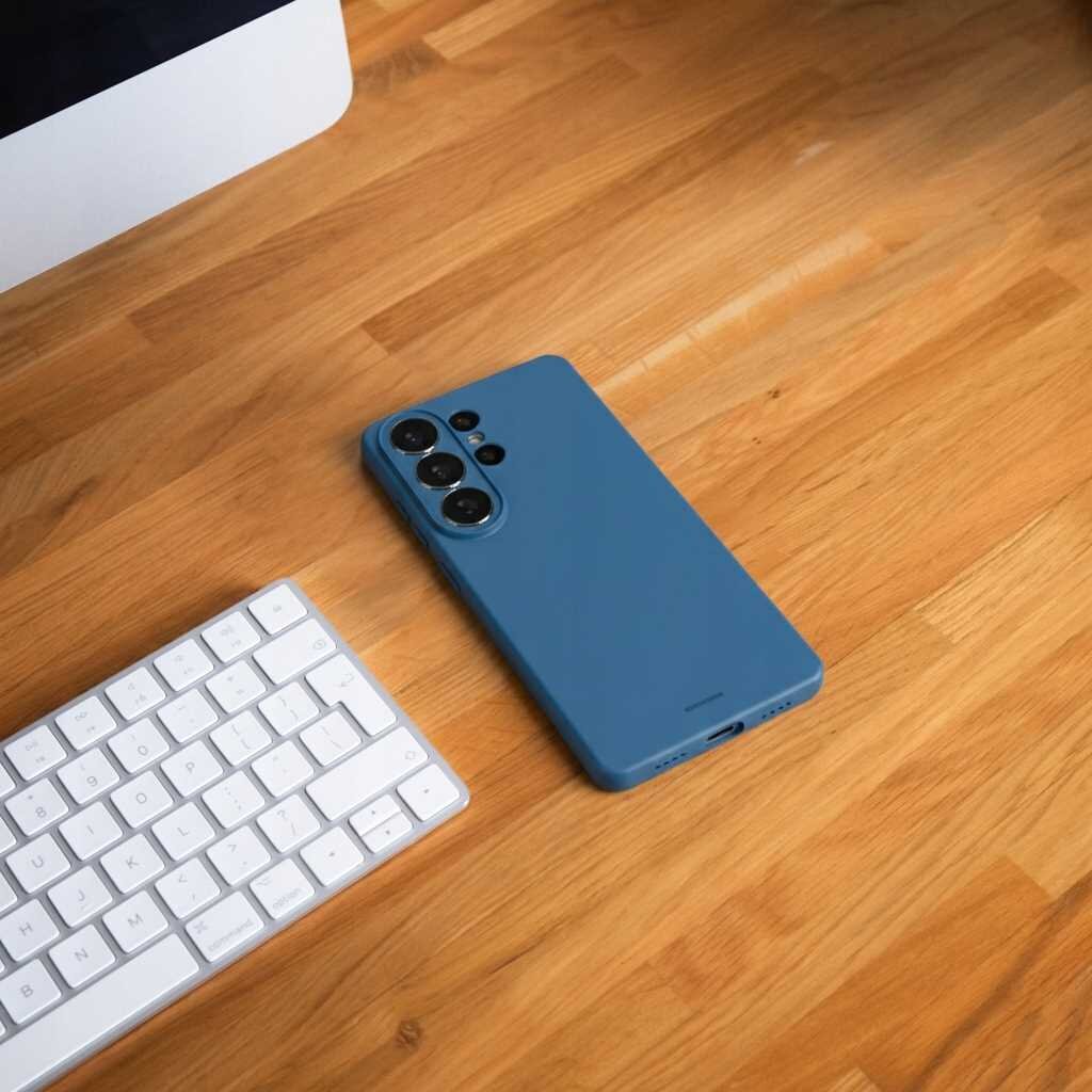 Na drewnianym blacie leży niebieskie silikonowe etui na telefon z trzema widocznymi obiektywami aparatu. Obok znajduje się biała klawiatura komputerowa.