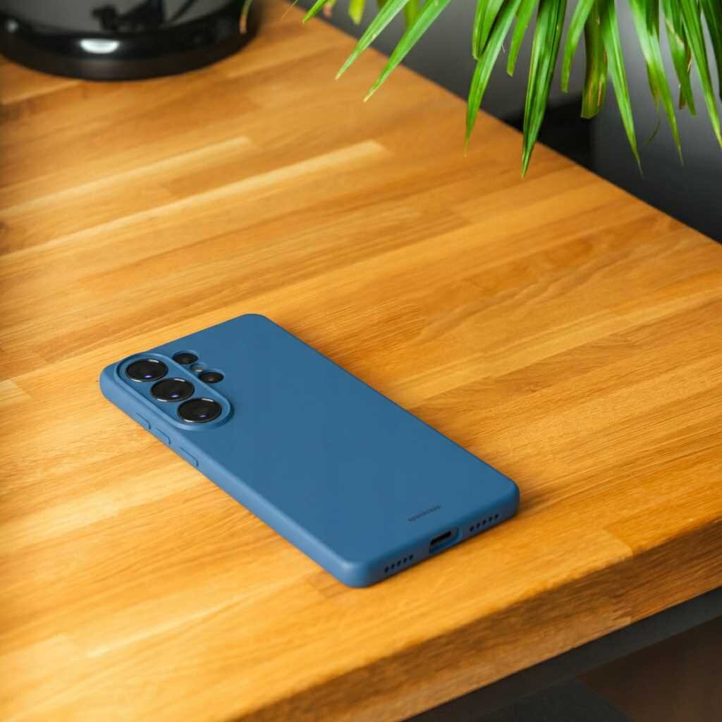 Na drewnianym blacie leży niebieskie silikonowe etui na telefon z trzema dużymi otworami na aparaty. W tle widoczne są zielone liście rośliny.