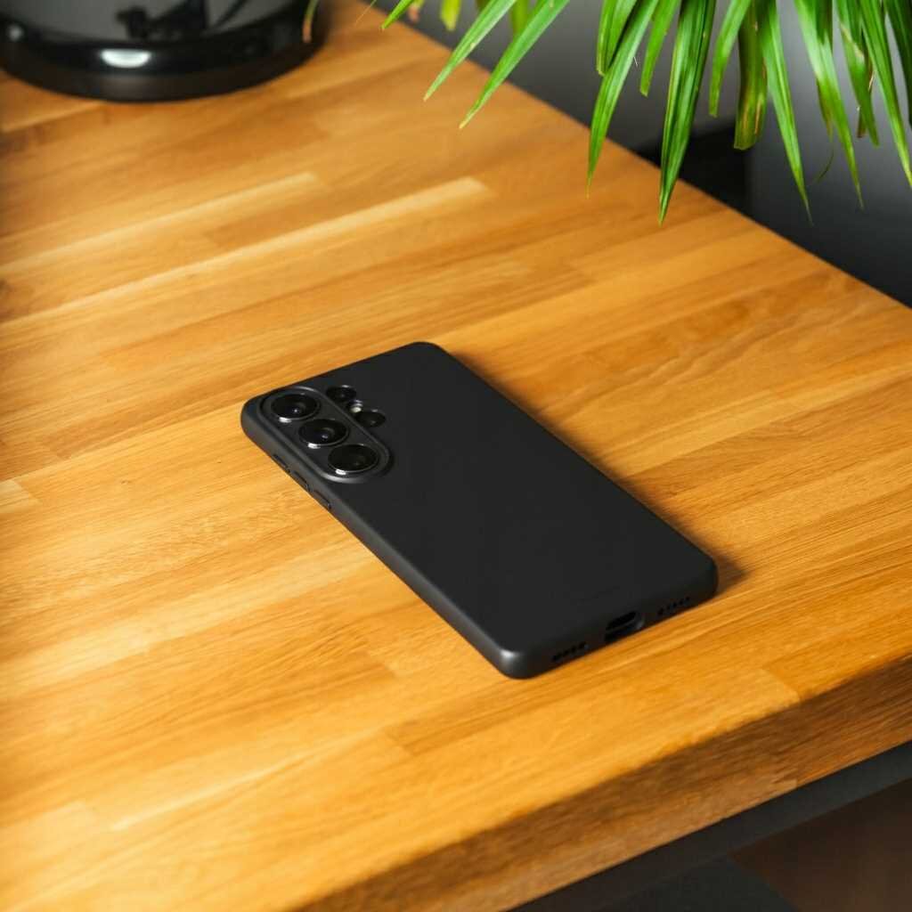 Na drewnianym blacie leży czarne silikonowe etui na smartfon z trzema widocznymi obiektywami aparatu. W tle widać fragment zielonej rośliny doniczkowej.