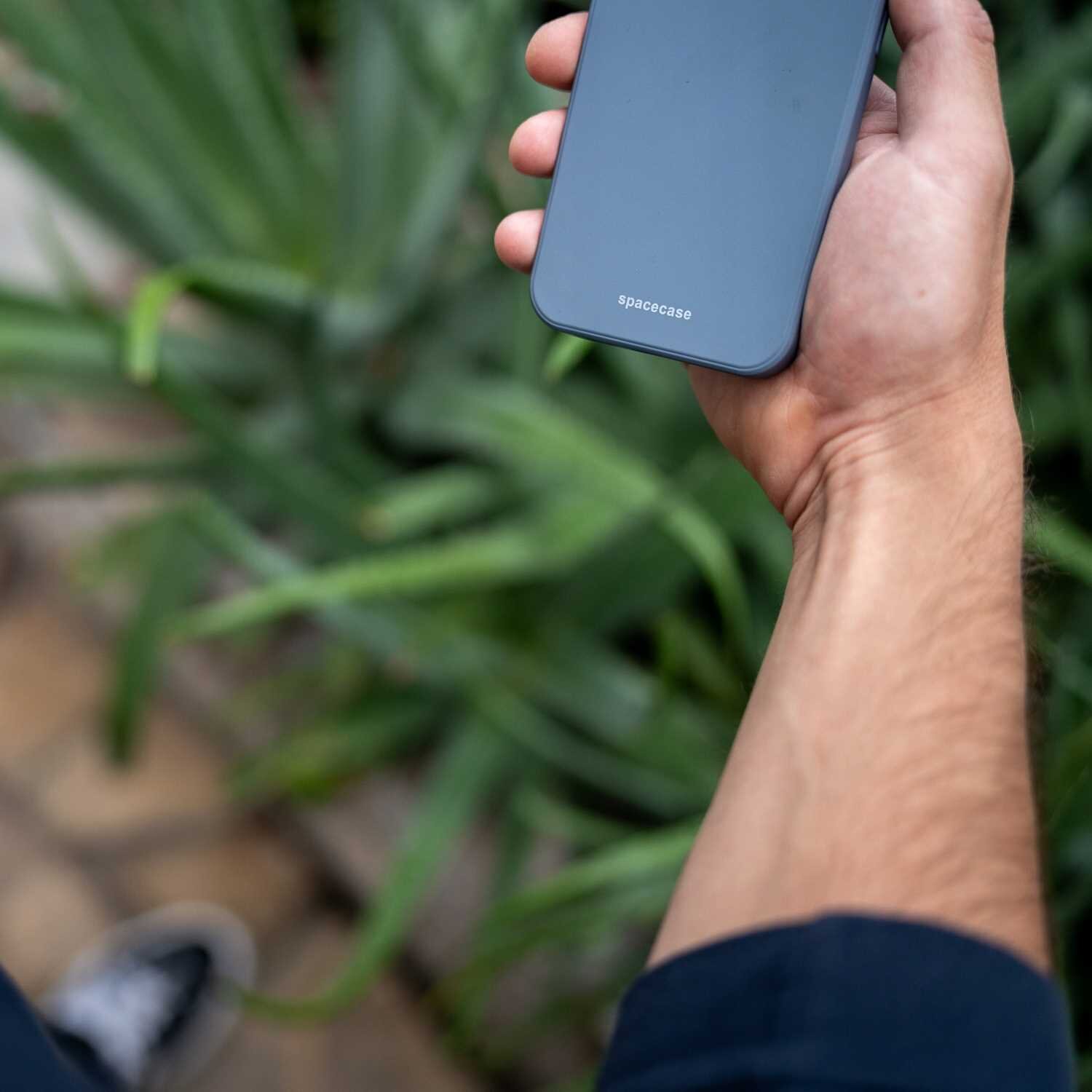 Osoba trzyma w dłoni telefon w niebieskim etui z napisem \'spacecase\'. W tle widoczne są zielone rośliny.