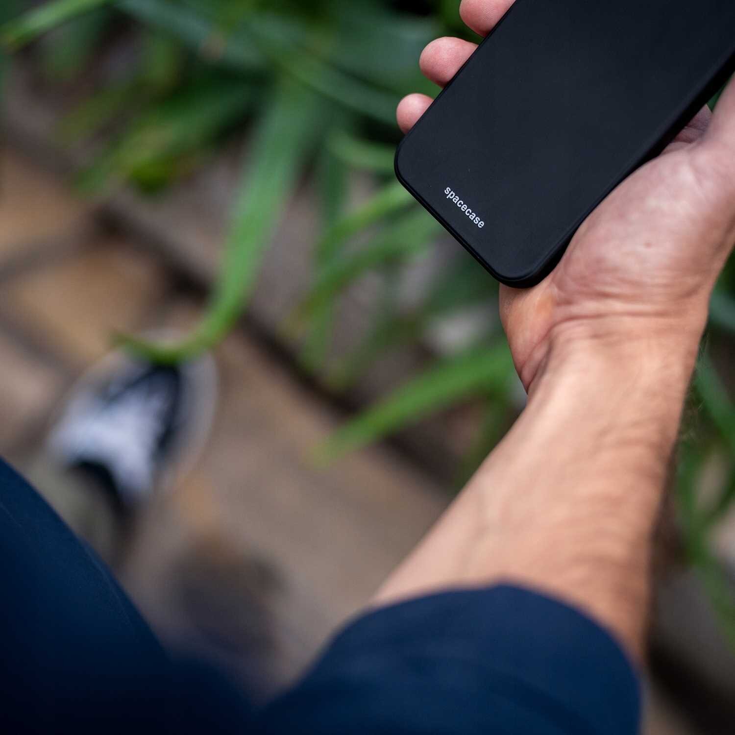 Osoba trzyma czarne silikonowe etui na telefon z widocznym napisem \'spacecase\'. W tle znajdują się zielone rośliny i fragment podłogi.