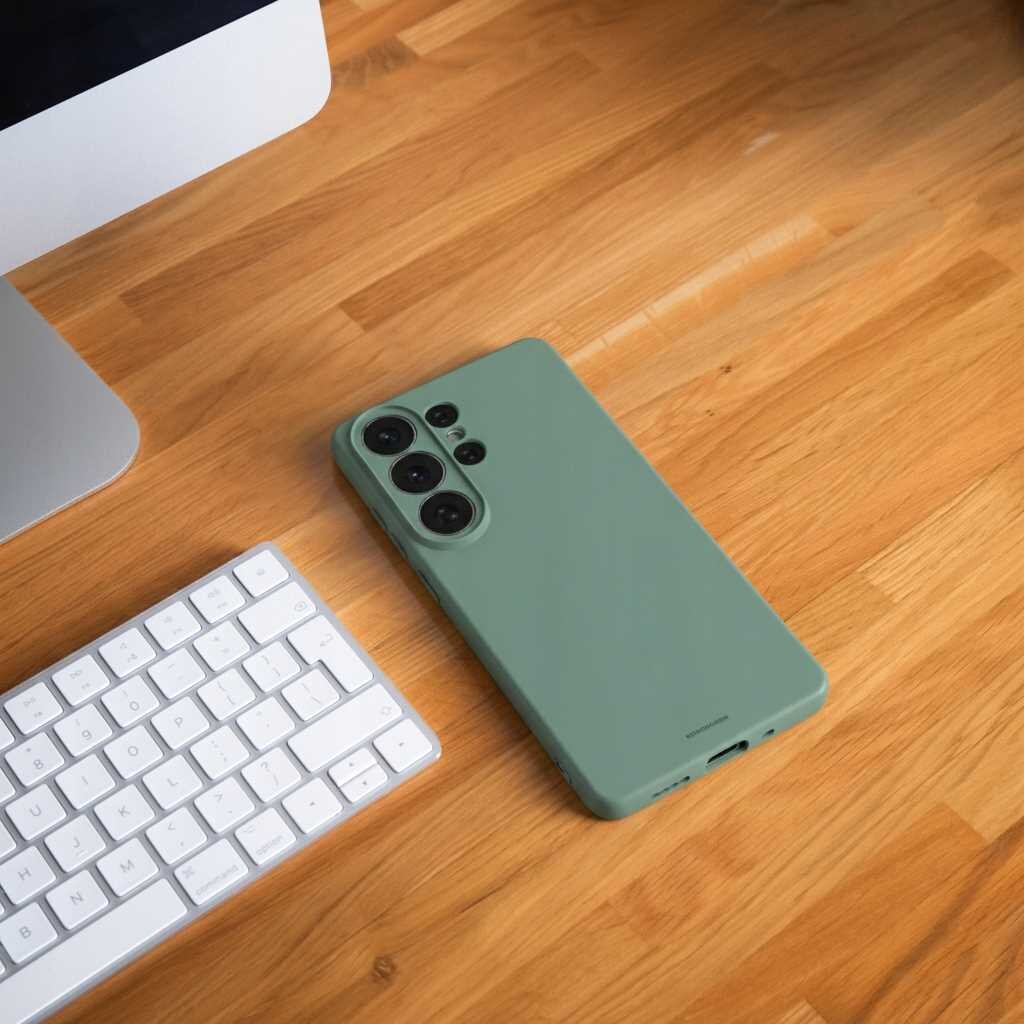 Na drewnianym blacie leży zielone silikonowe etui na smartfon z trzema widocznymi obiektywami aparatu. Obok znajduje się biała klawiatura i fragment monitora.