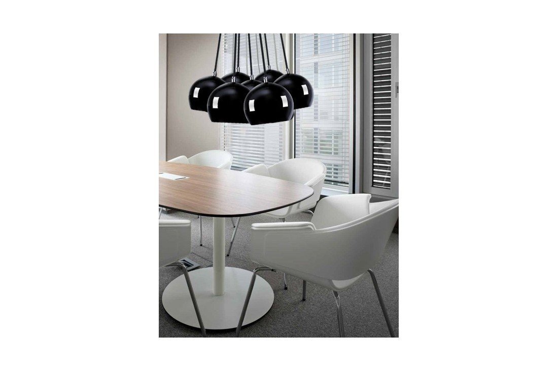 Na obrazie widoczna jest elegancka lampa wisząca z kilkoma czarnymi kloszami nad owalnym stołem w nowoczesnym wnętrzu. Wokół stołu ustawione są białe krzesła, a w tle znajdują się duże okna z żaluzjami.