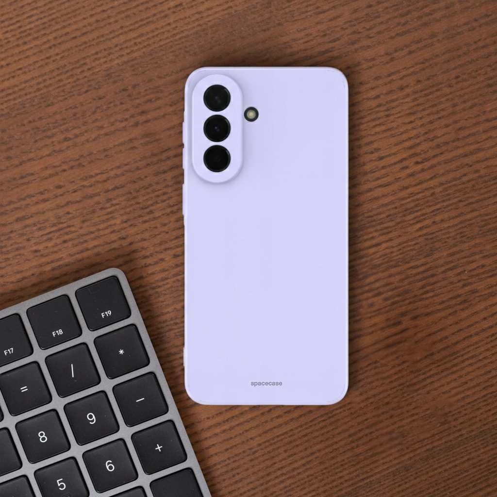 Na drewnianym stole leży fioletowe silikonowe etui na telefon z trzema otworami na aparaty i napisem \'spacecase\'. Obok znajduje się fragment klawiatury numerycznej.