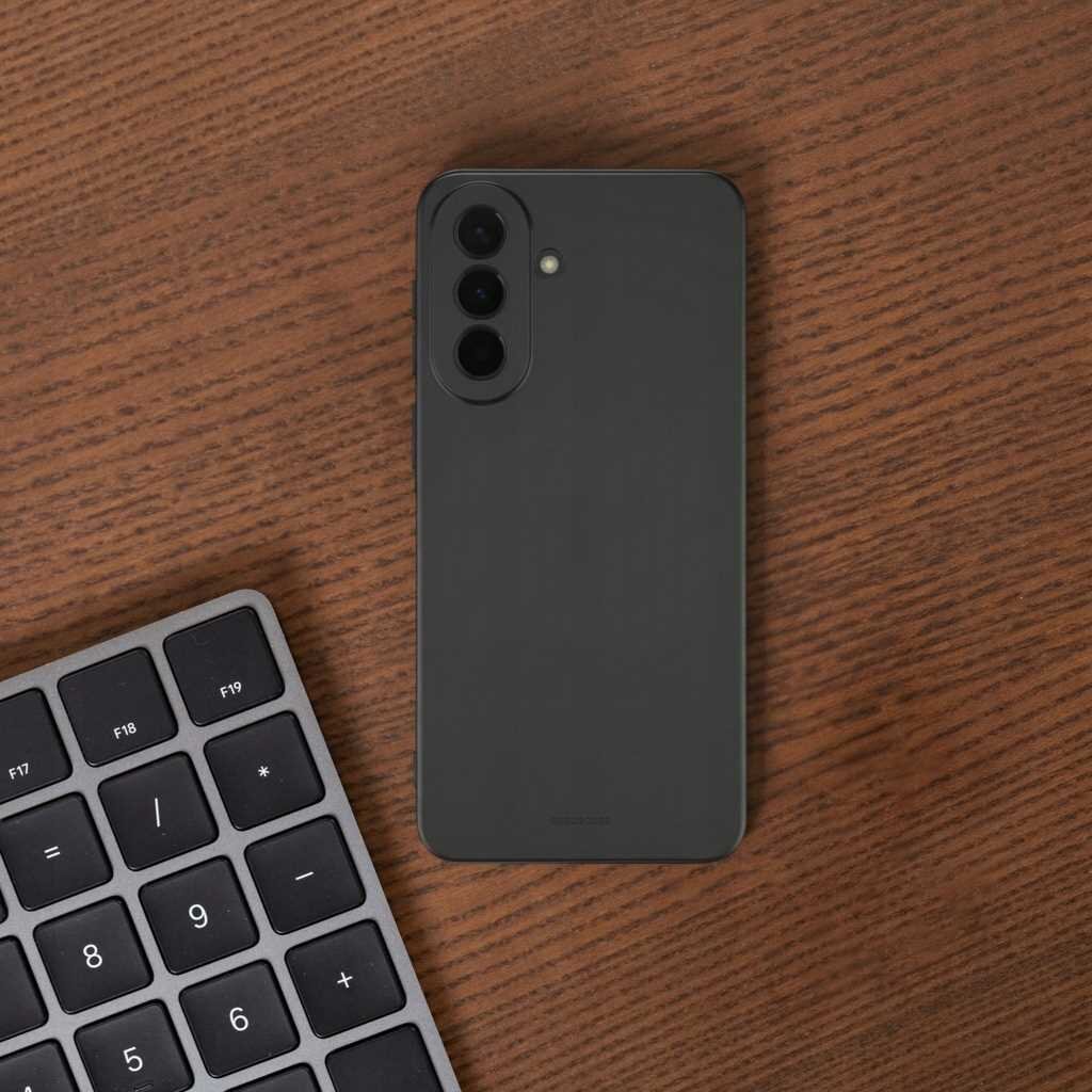 Na drewnianym blacie leży czarne silikonowe etui na telefon z trzema otworami na aparaty. Obok znajduje się fragment klawiatury z widocznymi klawiszami numerycznymi i funkcyjnymi.