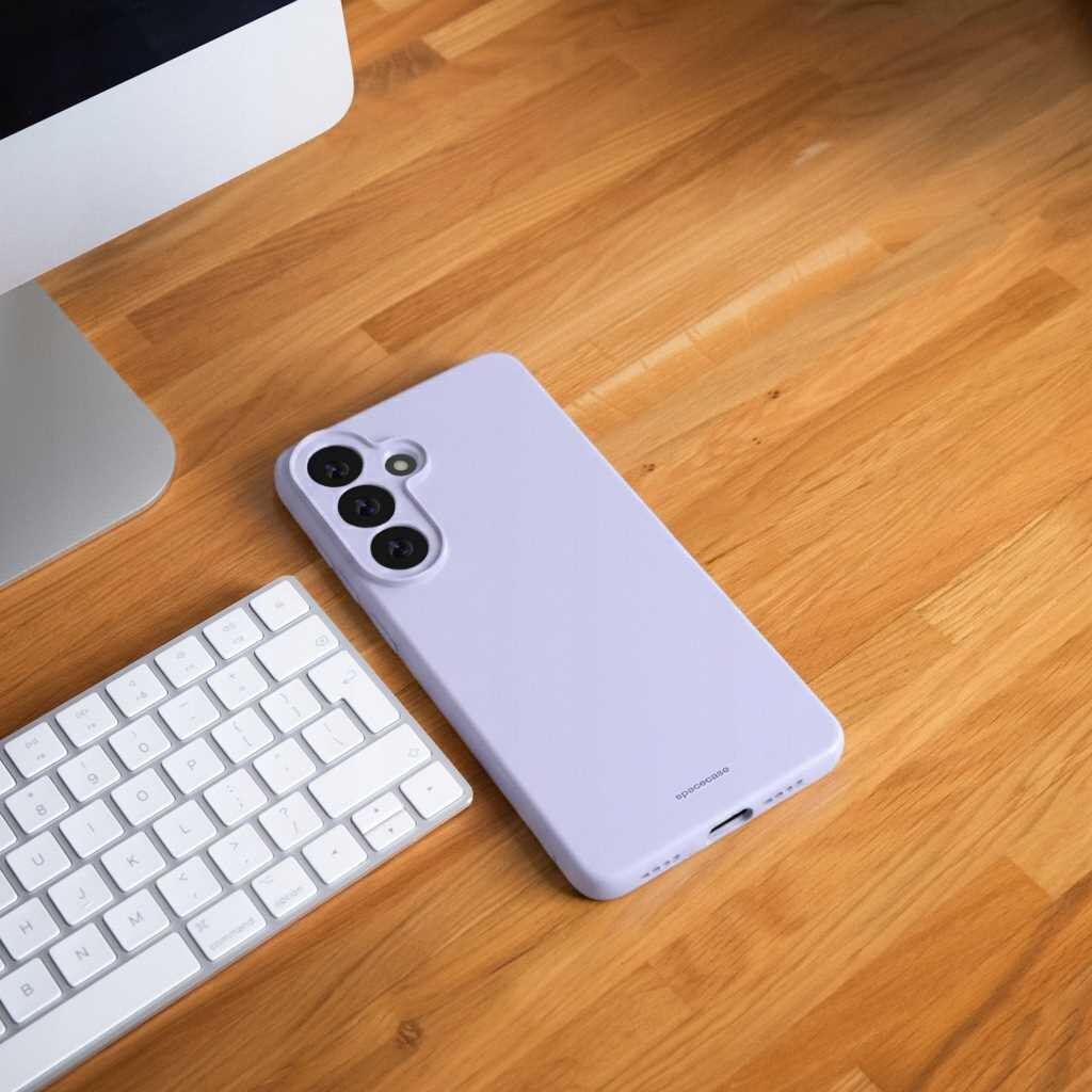 Na drewnianym blacie znajduje się fioletowe silikonowe etui na telefon z trzema otworami na aparaty. Obok leży biała klawiatura i fragment monitora.