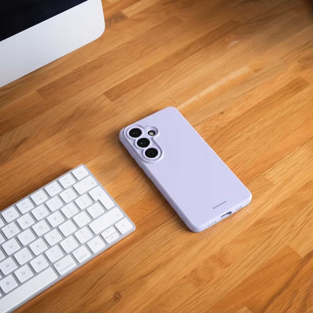 Na drewnianym biurku leży fioletowe silikonowe etui na telefon z trzema otworami na aparaty. Obok znajduje się biała klawiatura i fragment monitora.
