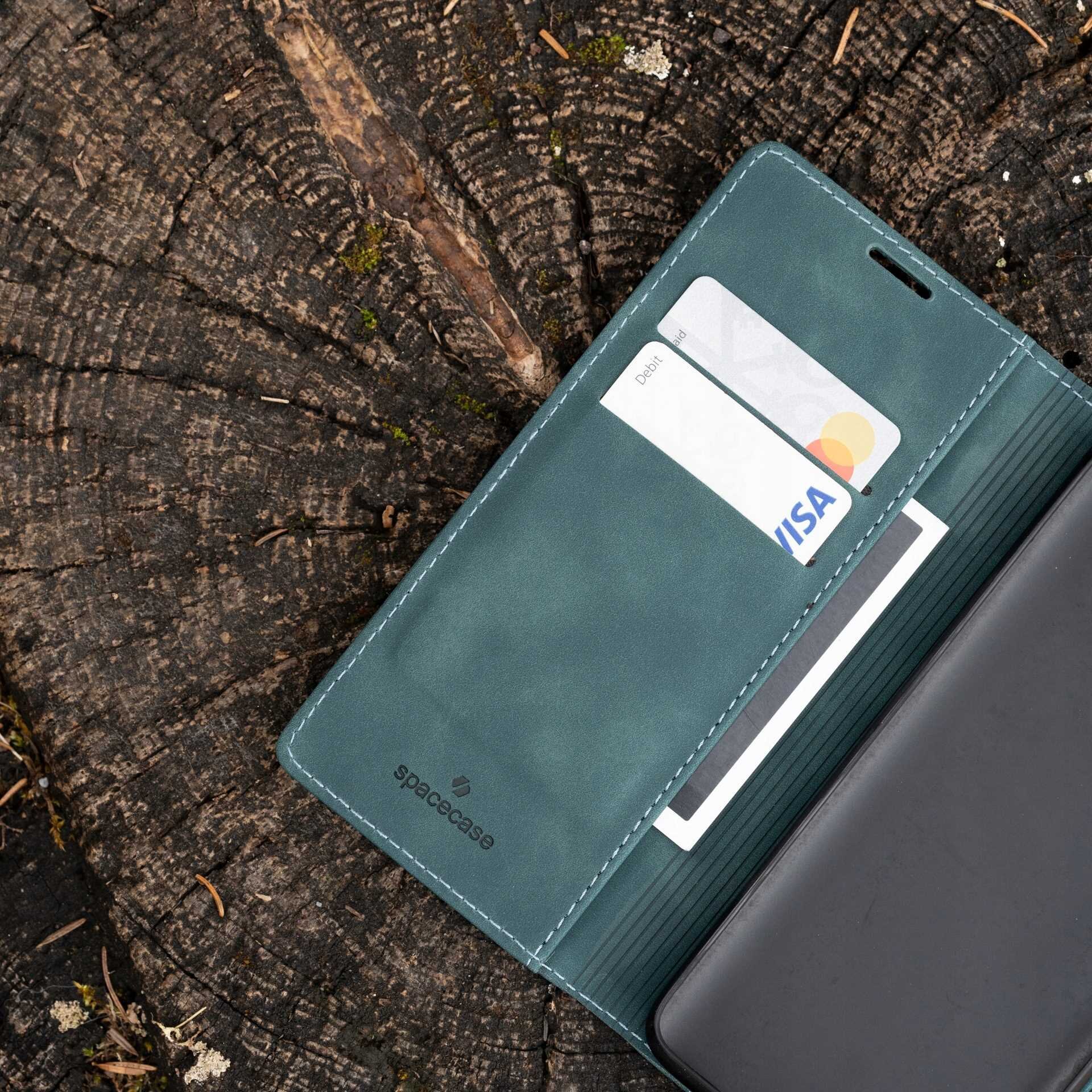 Niebieskie etui z klapką SPACECASE Wallet leży na drewnianym pniu, z widocznymi kieszeniami na karty, w tym kartą Visa. Na etui widoczne jest logo \'spacecase\'.