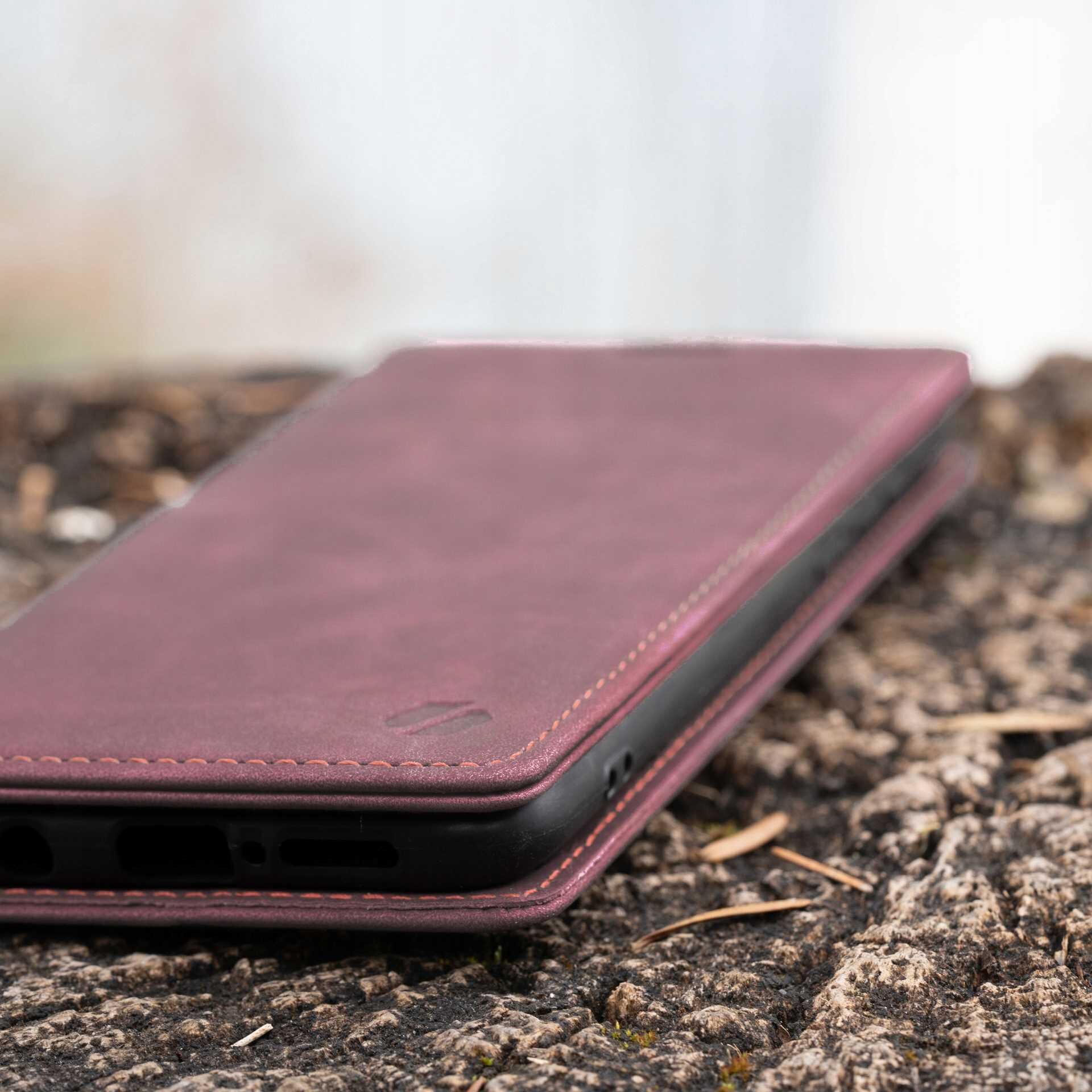 Czerwone etui z klapką leży na powierzchni przypominającej korę drzewa. Etui ma widoczne przeszycia i otwory na porty urządzenia.