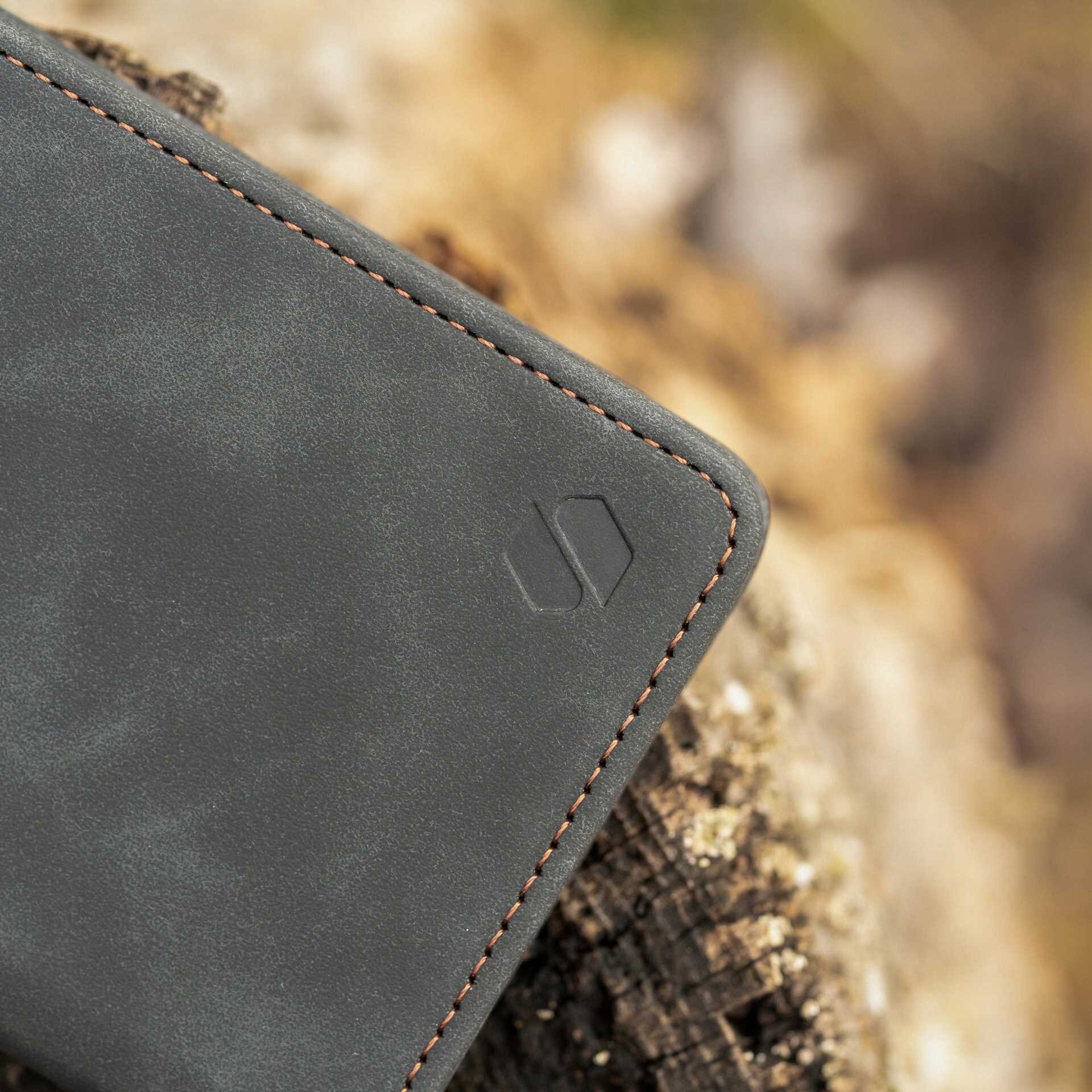 Czarny materiałowy etui z klapką z widocznym logo wytłoczonym na powierzchni oraz brązowym przeszyciem wzdłuż krawędzi. Tło stanowi rozmyty obraz drewna.