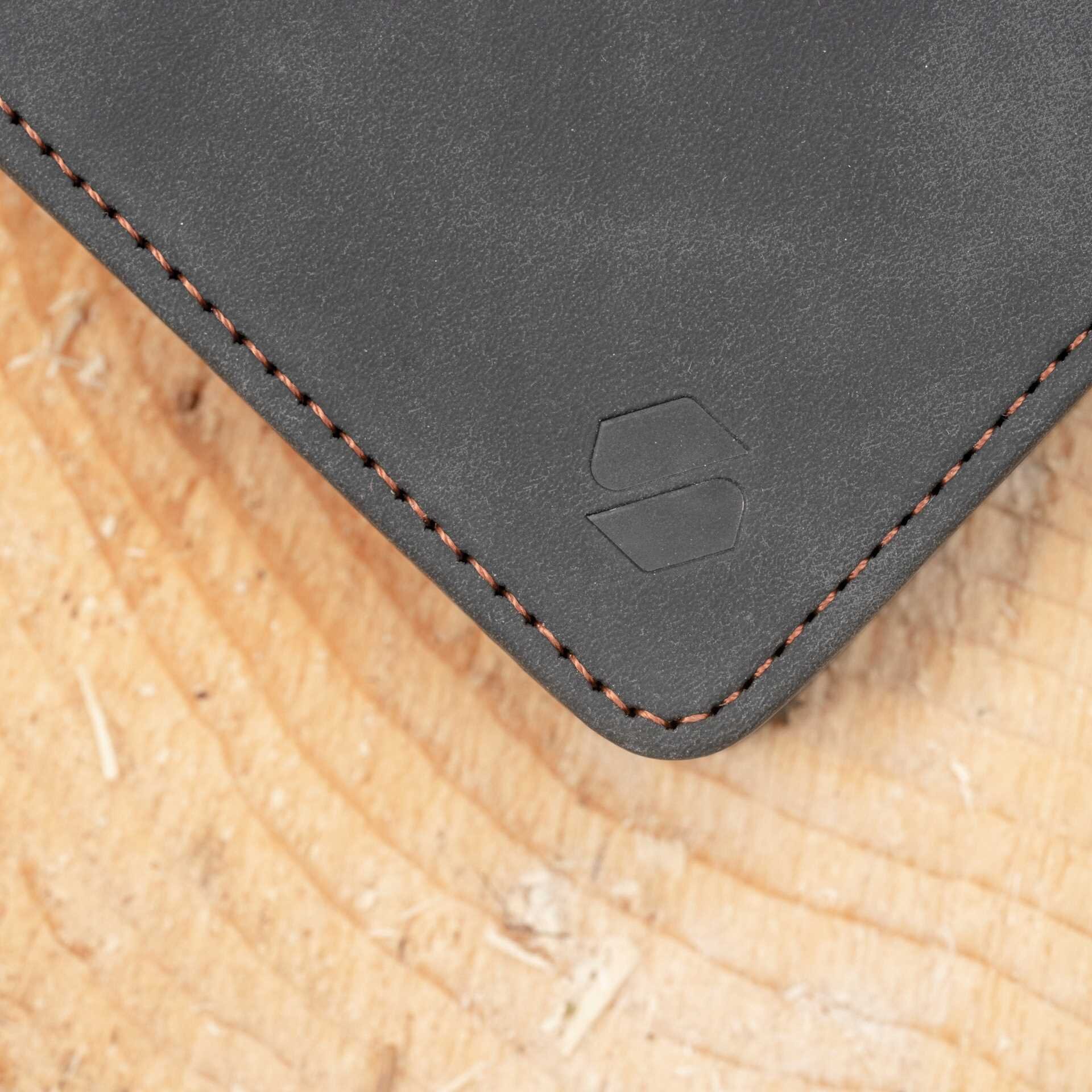 Czarny materiał z widocznym logo wytłoczonym w rogu oraz pomarańczowym przeszyciem na krawędzi. Tło to drewniana powierzchnia o jasnym odcieniu.