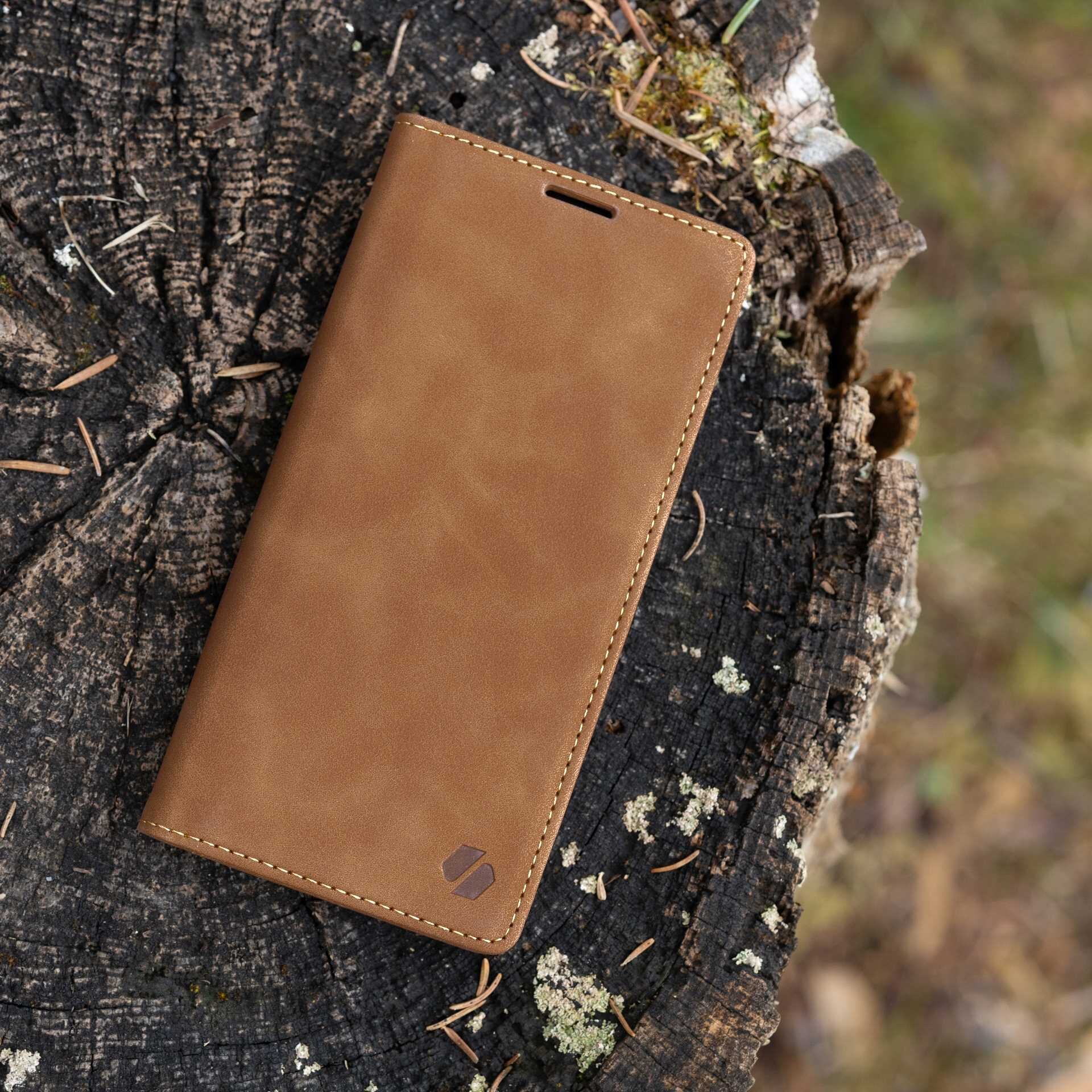 Brązowe etui z klapką leży na pniu drzewa. Na etui widoczne jest małe logo w dolnym rogu.