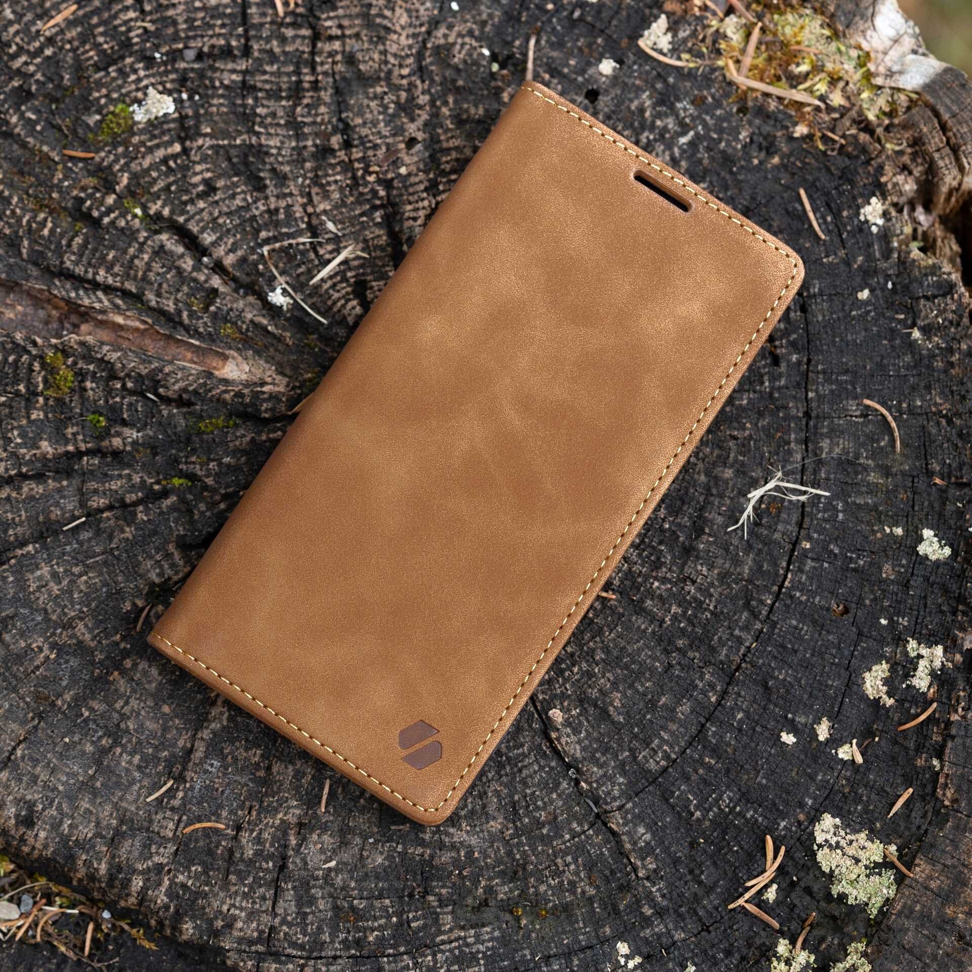 Brązowe etui z klapką leży na pniu drzewa. Na etui widoczne jest logo w formie dwóch połączonych kształtów.