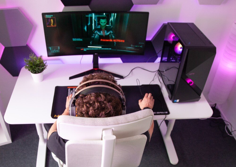 Komputer NTT Game Pro ZKG-I5145060-N01H Widok z góry na osobę w słuchawkach siedzącą na białym fotelu przed zakrzywionym monitorem, po prawej stronie stoi komputer