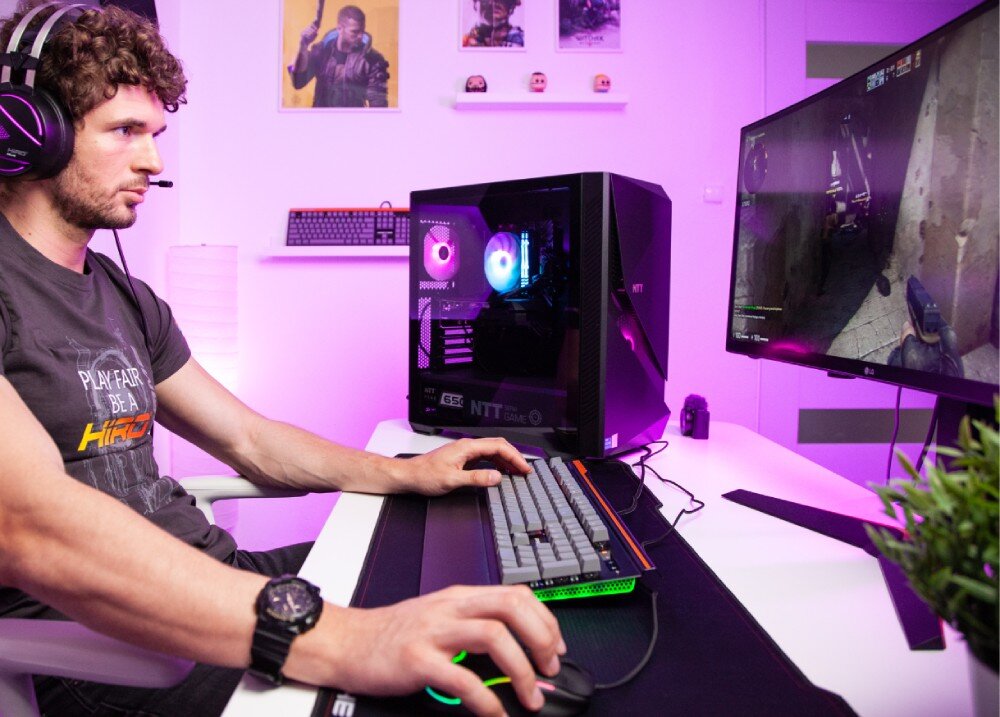 Komputer NTT Game Pro ZKG-I5145060-N01H Mężczyzna w słuchawkach gra na komputerze stacjonarnym, trzymając dłonie na podświetlanej klawiaturze i myszce