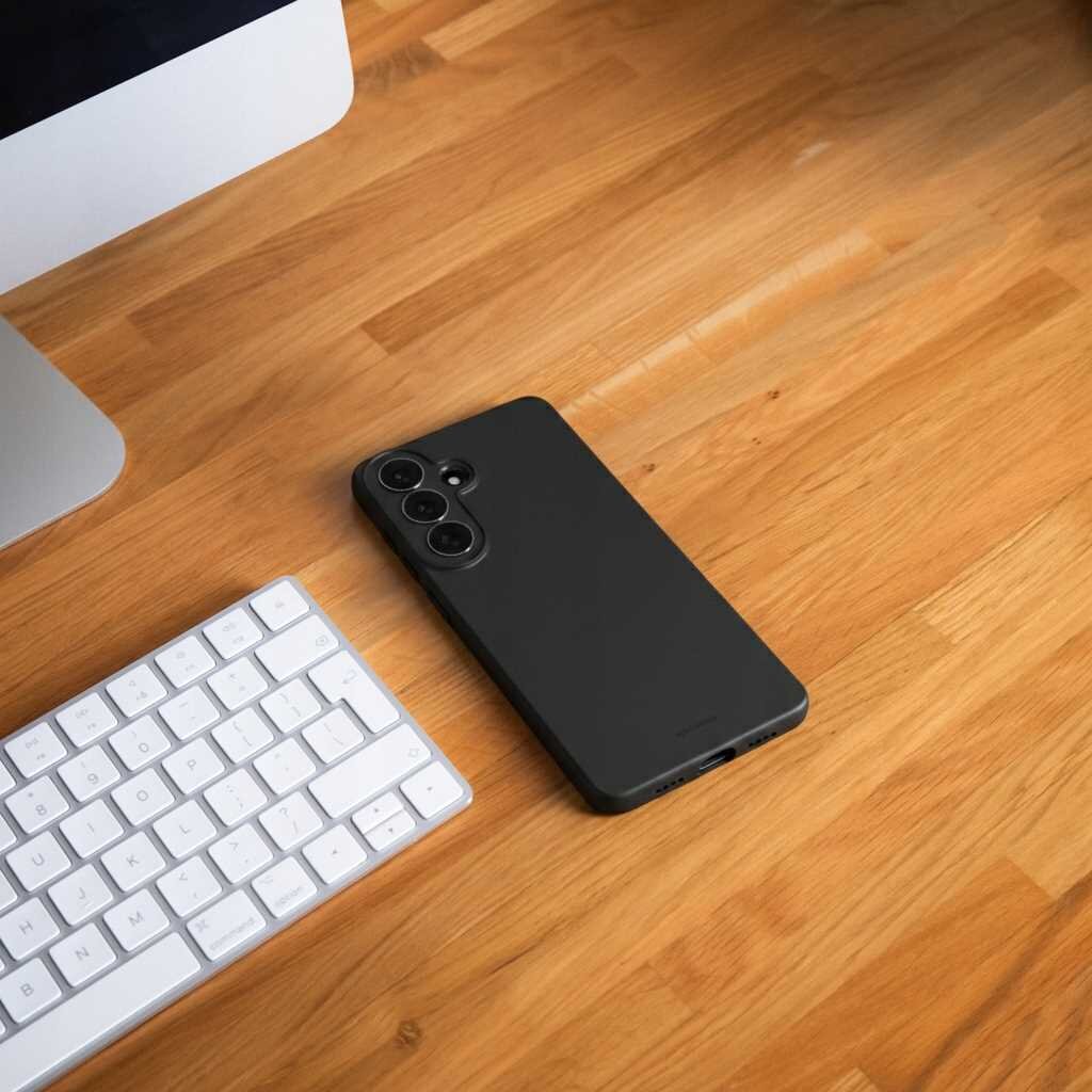 Na drewnianym biurku leży czarne silikonowe etui na smartfon z trzema otworami na aparaty. Obok znajduje się biała klawiatura i fragment monitora.