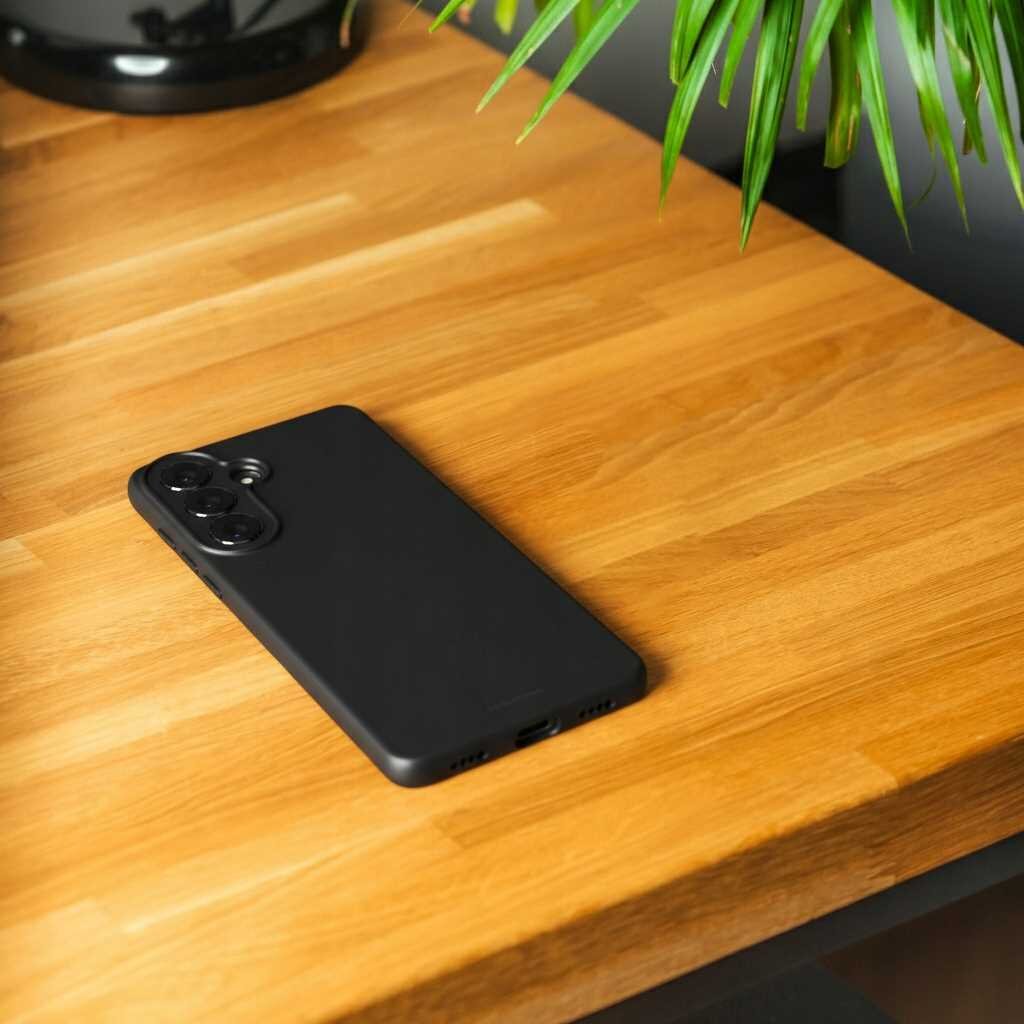 Czarne silikonowe etui na telefon leży na drewnianym blacie. W tle widoczne są zielone liście rośliny.