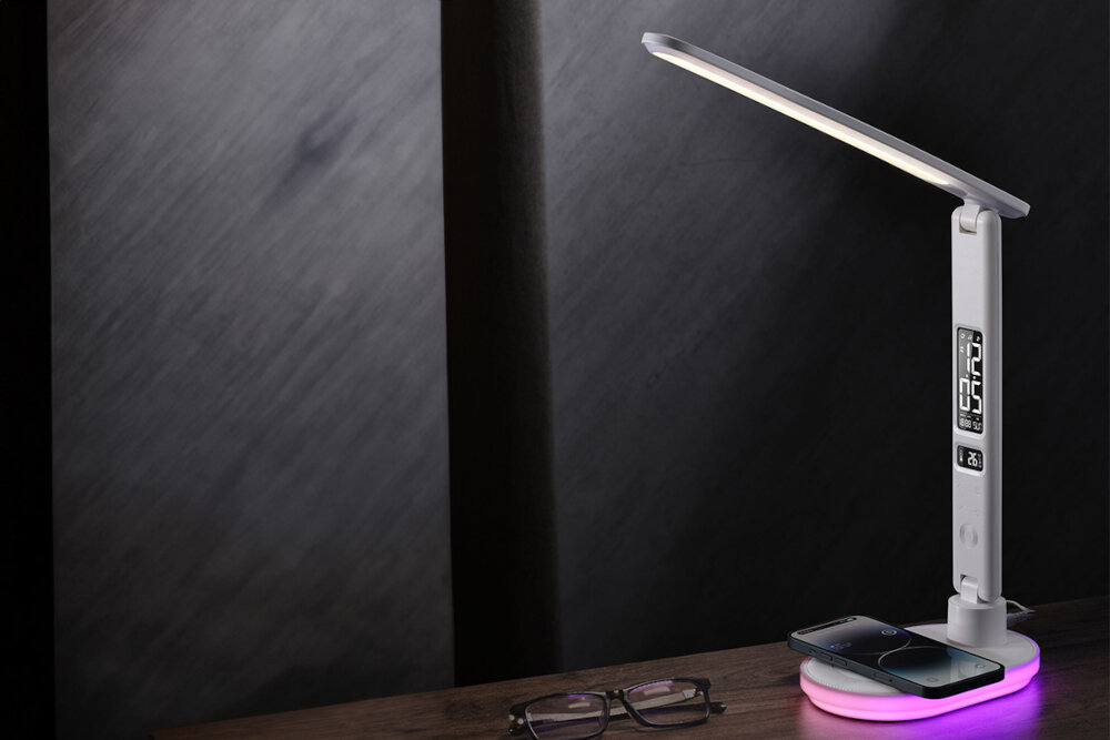 Na drewnianym blacie stoi nowoczesna lampka biurkowa z wyświetlaczem cyfrowym, obok której leżą okulary i smartfon. Podstawa lampki emituje delikatne, różowe światło.