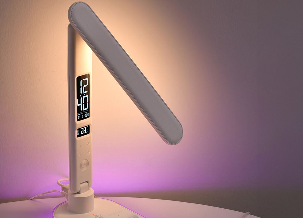 Lampka biurkowa z wyświetlaczem pokazującym godzinę 12:40 i temperaturę 28.3°C. Światło lampki jest skierowane na ścianę, tworząc fioletową poświatę.