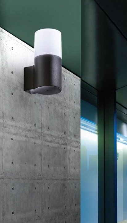 Na obrazie widoczny jest nowoczesny kinkiet zewnętrzny zamontowany na betonowej ścianie. Lampa ma cylindryczny kształt z ciemną podstawą i jasnym kloszem.