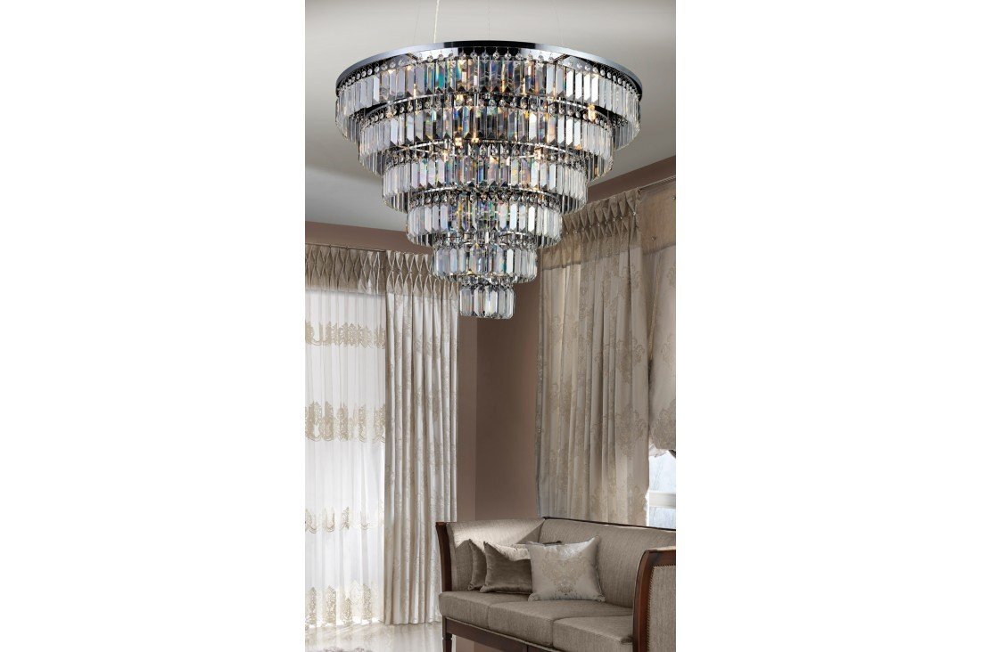 Na obrazie widoczna jest elegancka lampa wisząca złożona z wielu poziomów szklanych elementów, zawieszona w stylowym wnętrzu z zasłonami i sofą. Pomieszczenie jest urządzone w stonowanych kolorach, co podkreśla luksusowy charakter lampy.