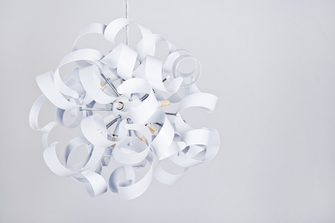 Lampa wisząca o nowoczesnym designie, składająca się z wielu białych, zakrzywionych elementów tworzących kulistą formę. W centrum widoczne są żarówki i metalowe elementy konstrukcyjne.