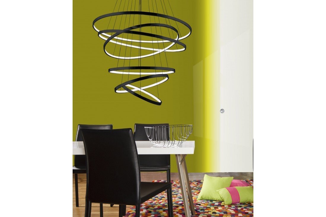 Na obrazie widoczna jest designerska lampa wisząca z kilkoma okrągłymi elementami świetlnymi, zawieszona nad stołem. W tle znajduje się zielona ściana, a na podłodze kolorowy dywan i poduszki.